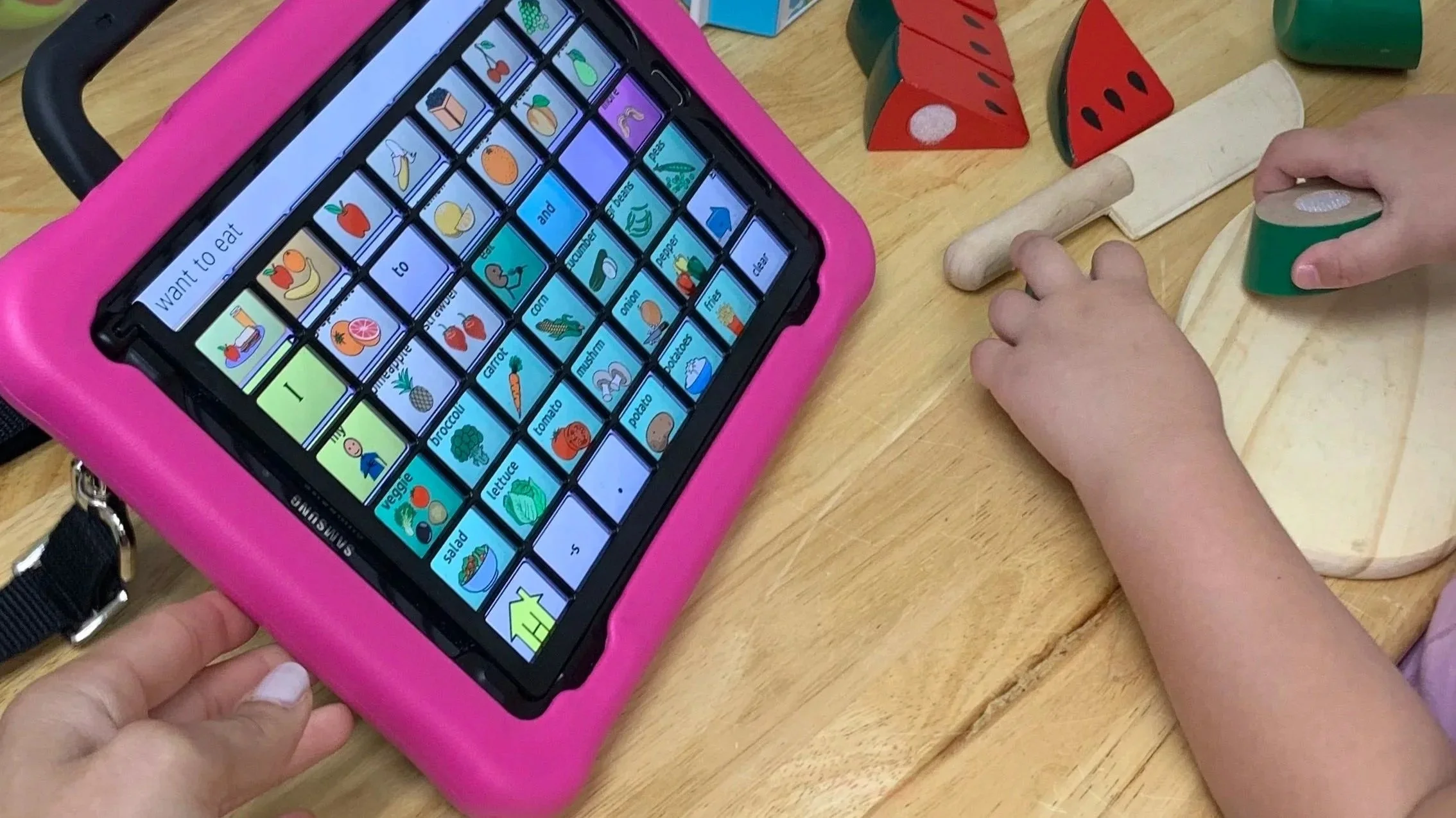 A child playing with a tablet and wooden toy food on a table.