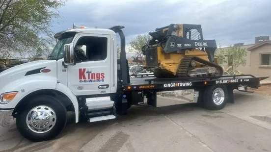 Tow truck carrying a mini excavator on a parking lot, with trees and buildings in the background.