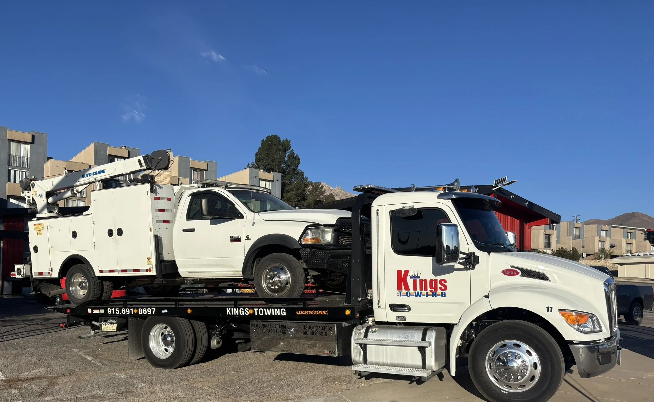White tow truck labeled 'Kings Towing' transporting a white utility truck with a crane in a parking lot with apartment buildings and mountains in the background.