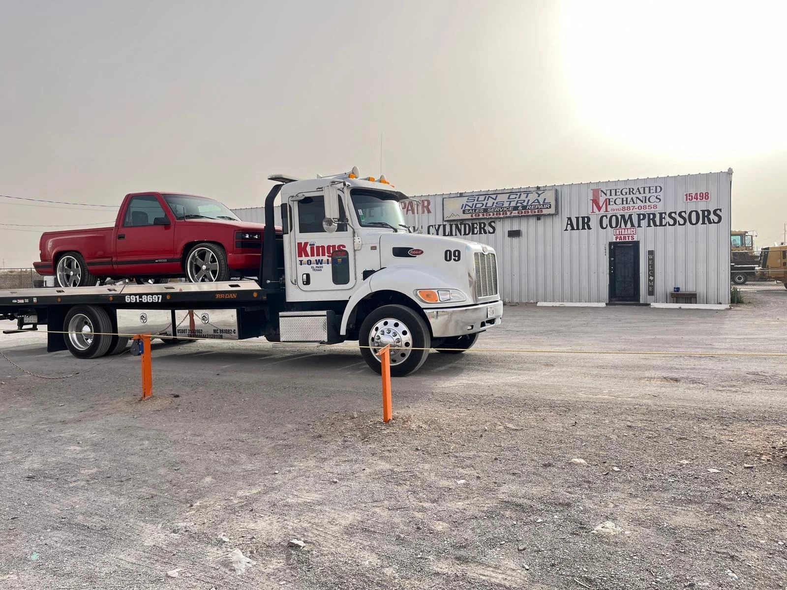 A white tow truck carrying a red pickup truck is parked outside an auto repair shop called Kings Towing. The shop specializes in air compressors, cylinders, and industrial repairs.