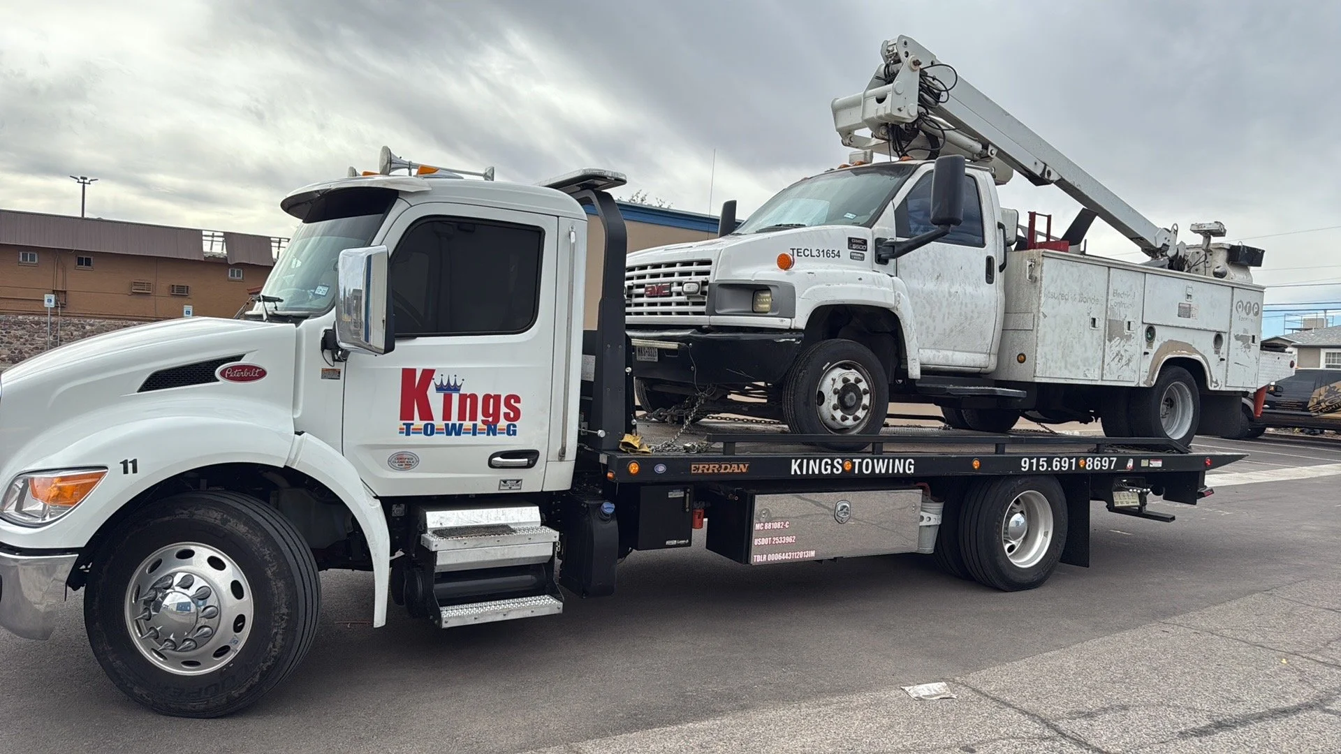 A white tow truck with the logo 'Kings Towing' on the door is parked on the street, carrying a white utility truck with a crane on top.