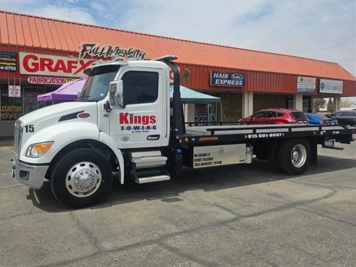 A white tow truck with a red and black logo that says 'Kings Towing' is parked in front of a strip mall. The truck is carrying two cars, a red one and a blue one, on its flatbed.
