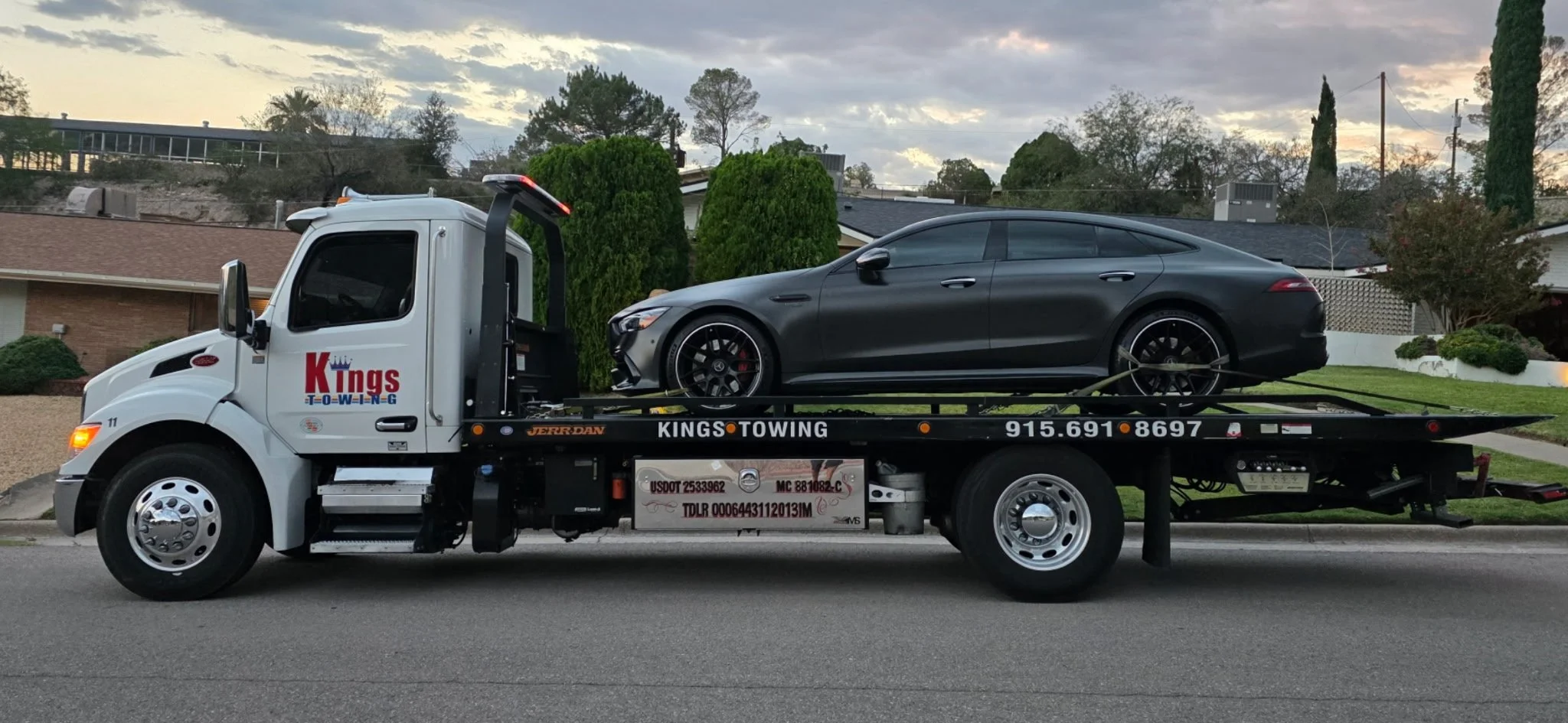 A tow truck hauling a black sports car on a residential street with houses and trees in the background during sunset.