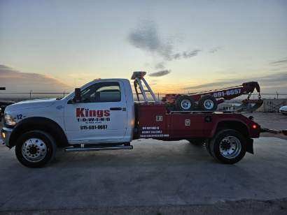 A tow truck with a white cab and red bed, carrying a small car on a flatbed, parked on a sandy surface during sunset.