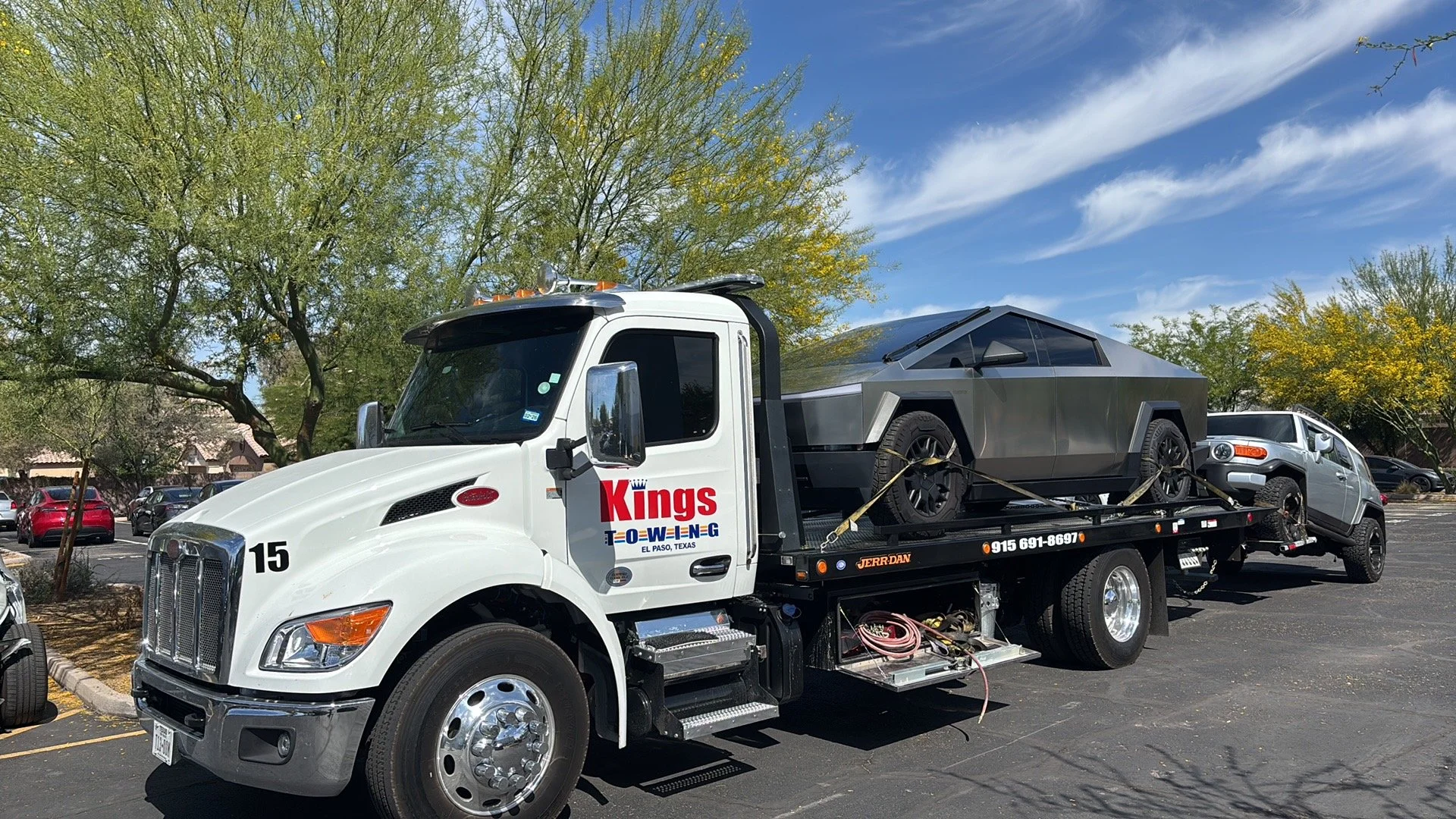 A tow truck carrying a futuristic, angular black vehicle in a parking lot with trees and other cars in the background.