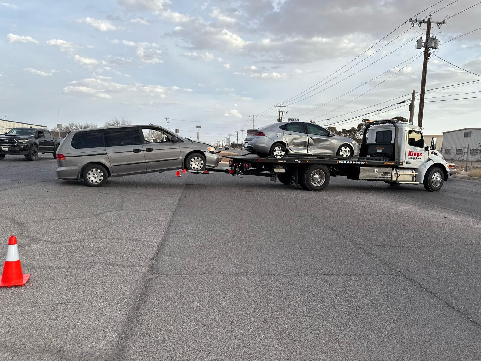 A tow truck is transporting a silver sedan which collided with a dark gray minivan. The minivan is connected to the tow truck with a hook and chain, and the sedan is on the flatbed of the tow truck. There are orange safety cones placed around the sce