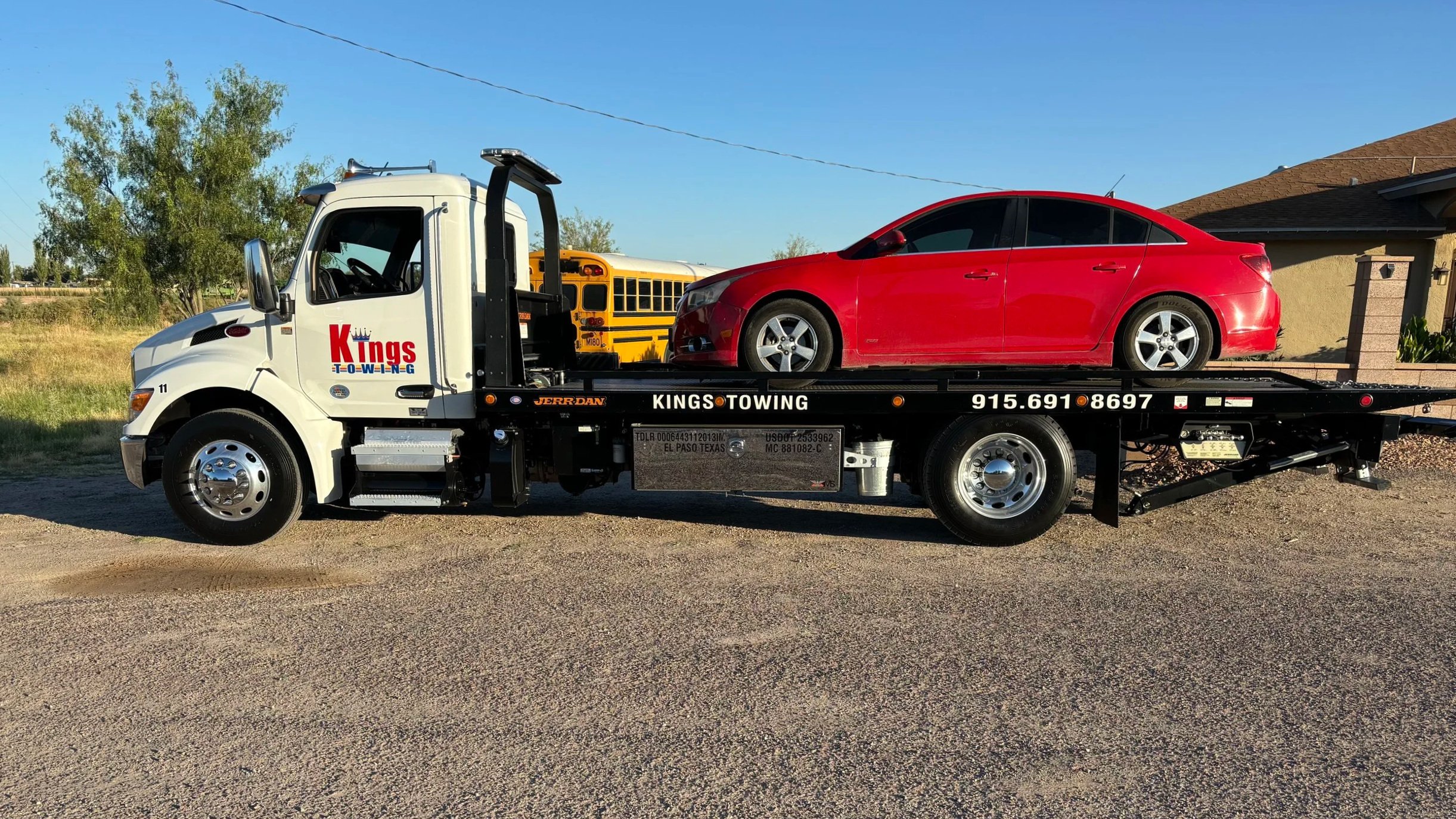 A white tow truck with 'Kings Towing' branding carrying a red sedan in a residential area with houses and trees in the background.