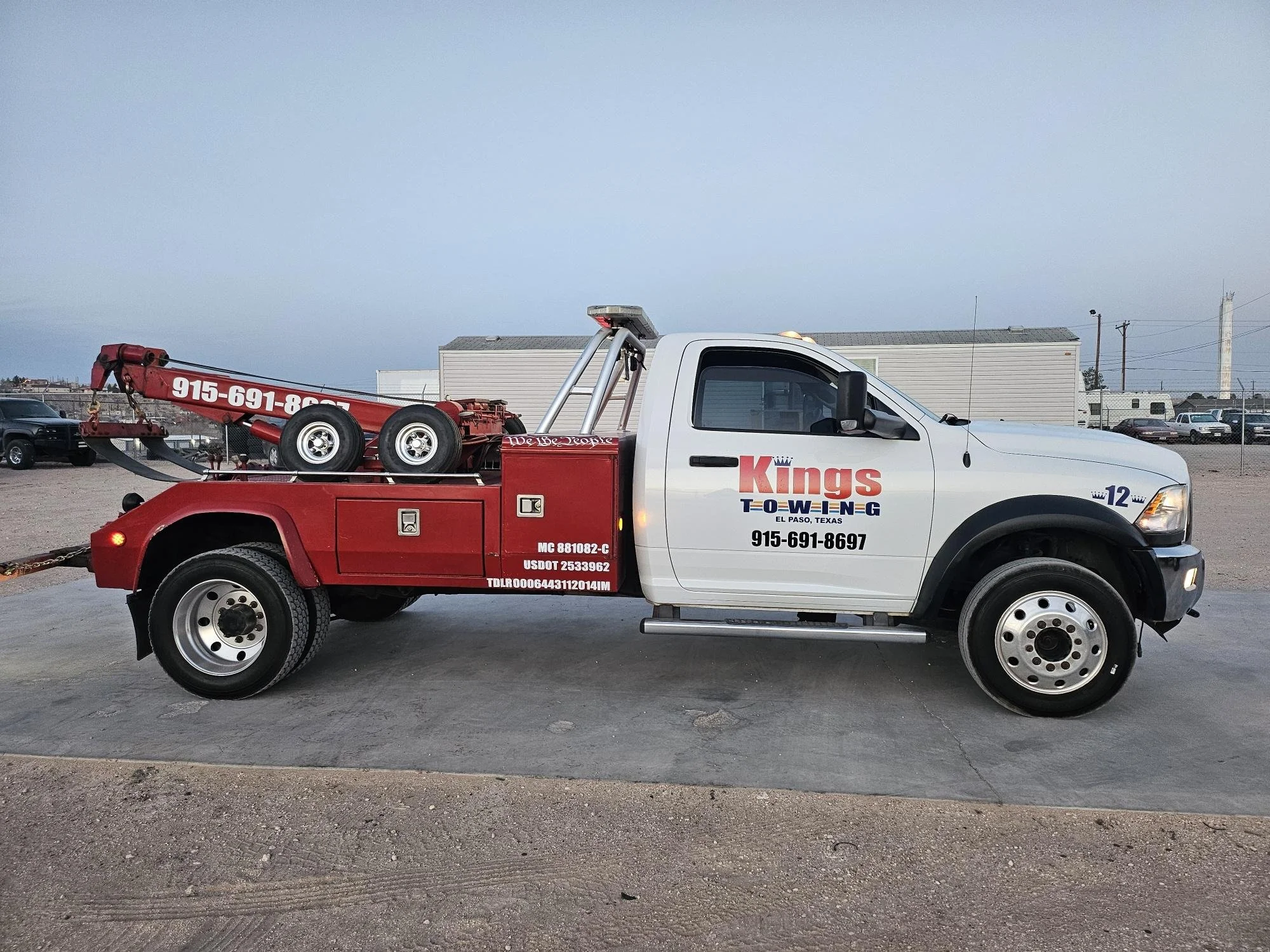 White truck with red towing equipment and company information, parked on a lot. Background includes a cloudy sky, industrial buildings, and other vehicles.