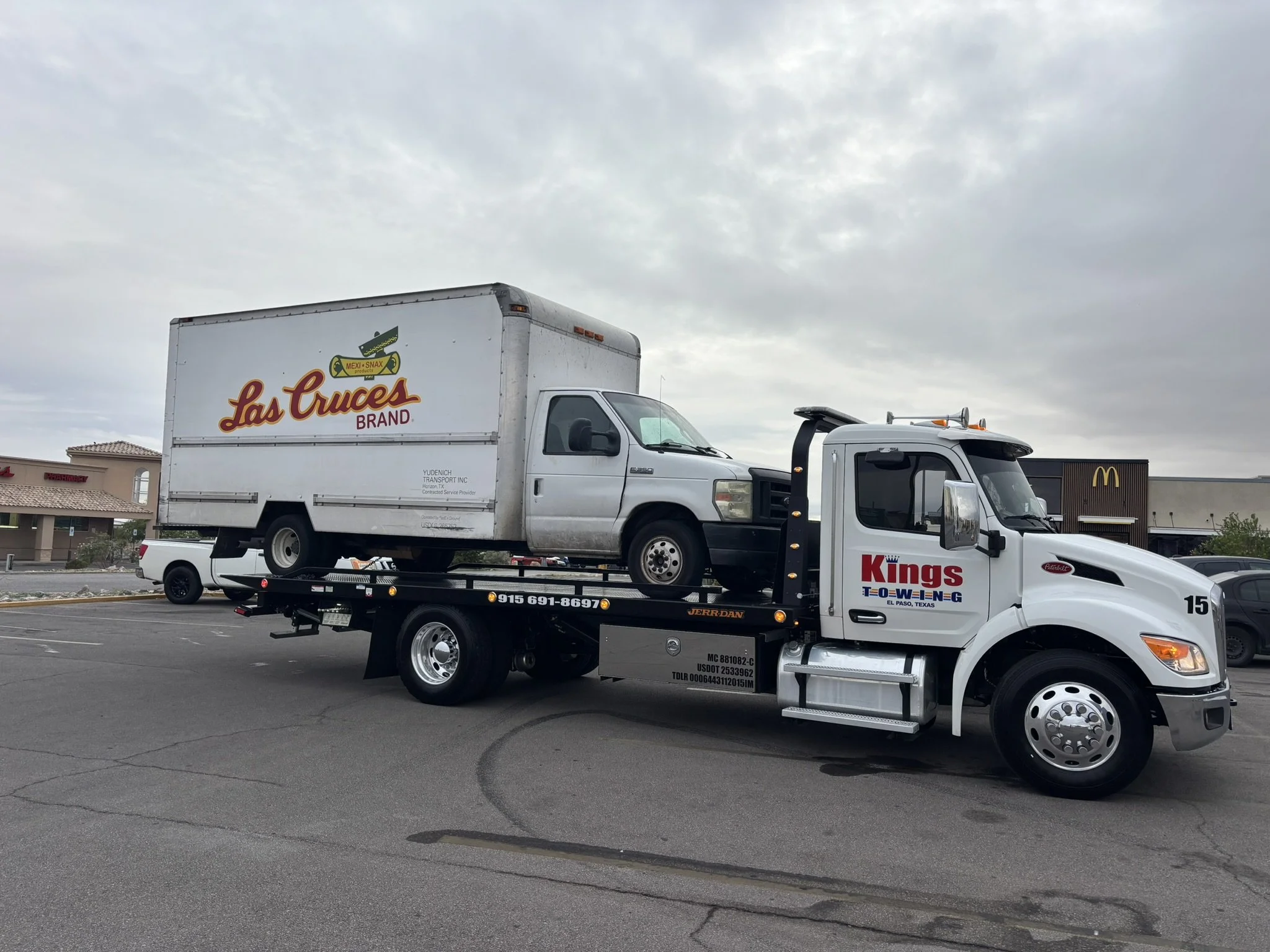 A tow truck is transporting a white box truck with Los Cruces branding on its side in a parking lot. The area has other vehicles and a McDonald's in the background under a cloudy sky.