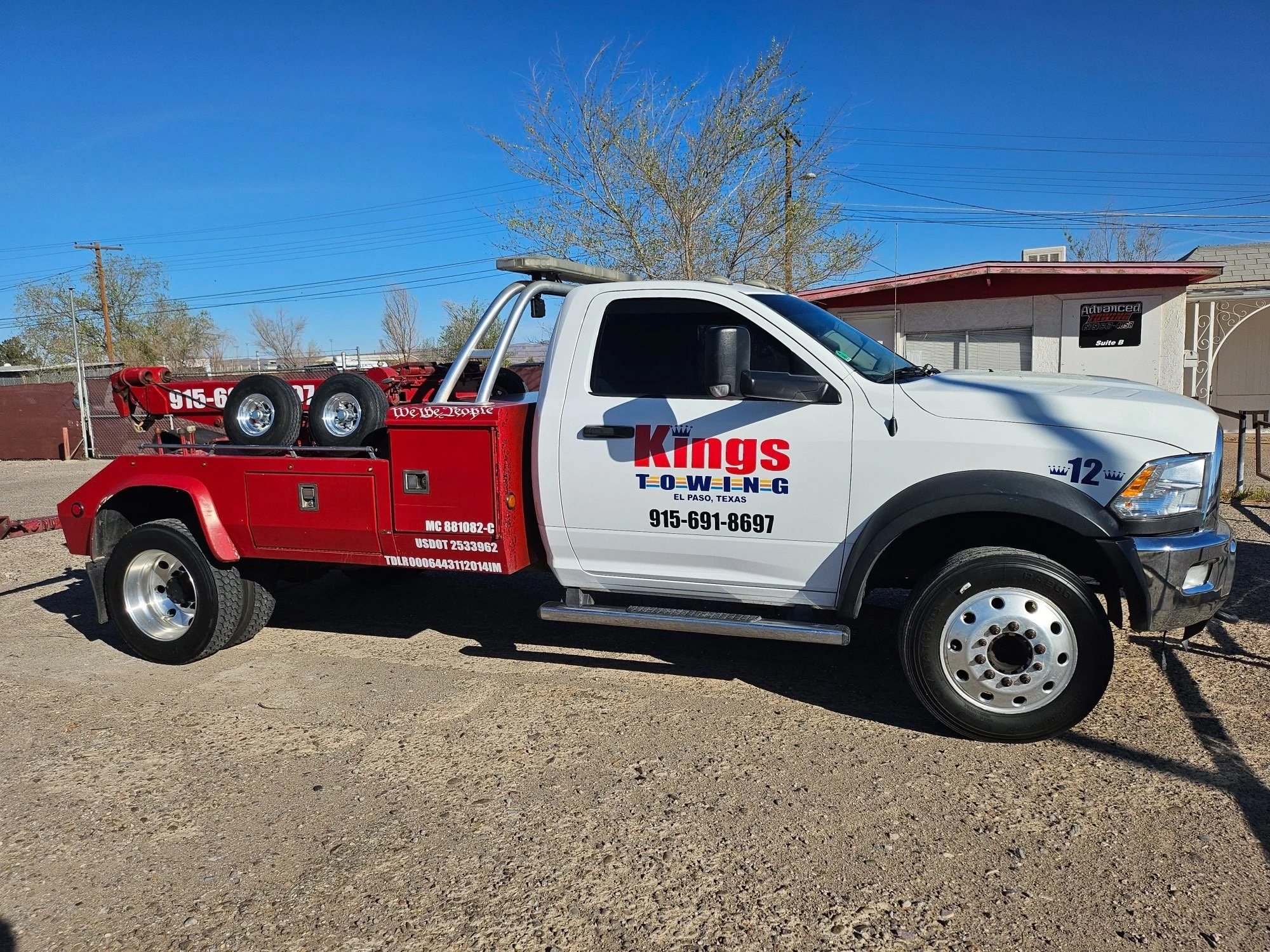A tow truck painted in white and red with company branding from Kings Towing in El Paso, Texas, parked on a gravel lot with a building and trees in the background.