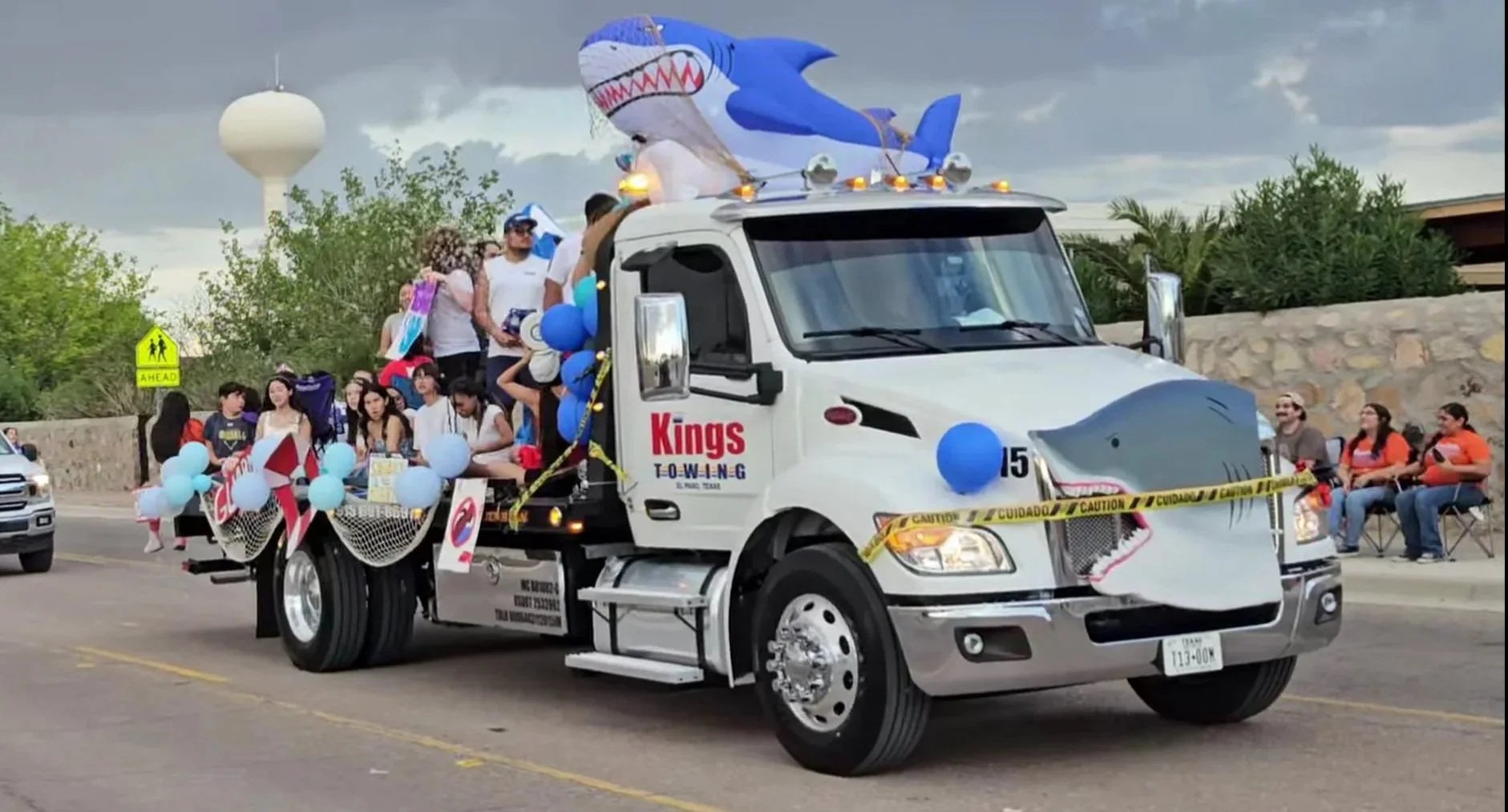 A decorated parade float designed to look like a shark, with people on and around it, in a parade on a city street with spectators sitting on the sidewalk, balloons, and balloons, a shark head on the front, and a large inflatable shark on top.