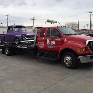 A red tow truck from Kings towing company carrying a purple vintage car on its flatbed in a parking lot.
