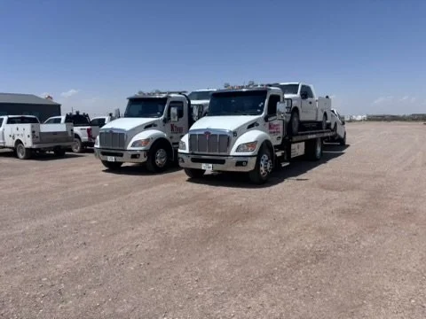 Two large white tow trucks with flatbed trailers parked on a dirt lot under clear blue sky.