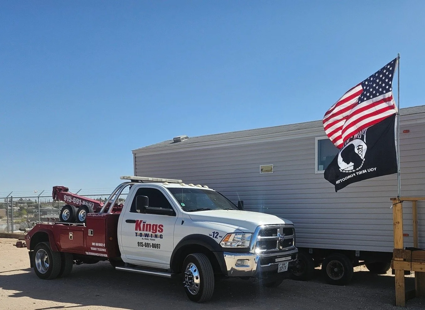 A white and red tow truck with the logo 'Kings Towing El Paso, Texas' parked next to a tiny house and flags, including an American flag and a black flag with a white emblem and text.