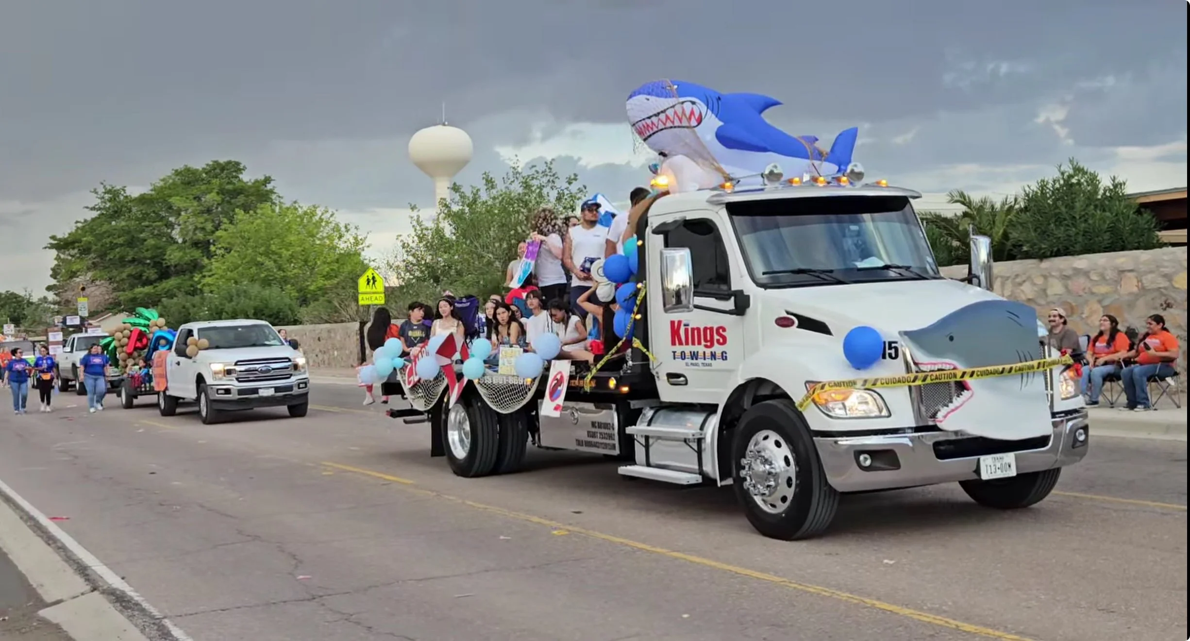 A parade float on a street with a large inflatable shark on the front of a truck decorated with balloons and strings of netting, surrounded by spectators and other vehicles, including another decorated truck behind it, under cloudy skies.