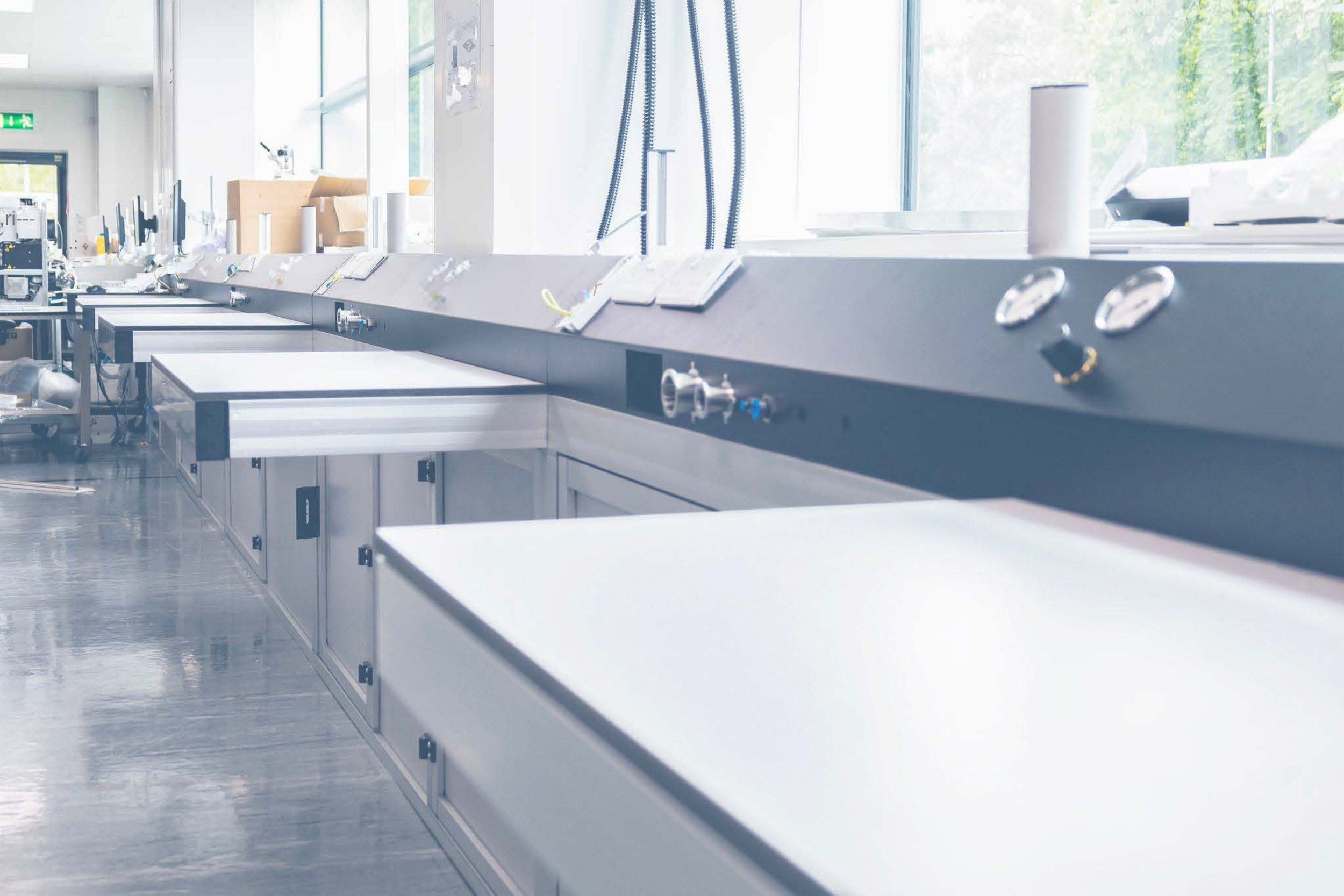 Empty stainless steel laboratory benches and equipment in a modern, well-lit laboratory with large windows showing green outdoor scenery.
