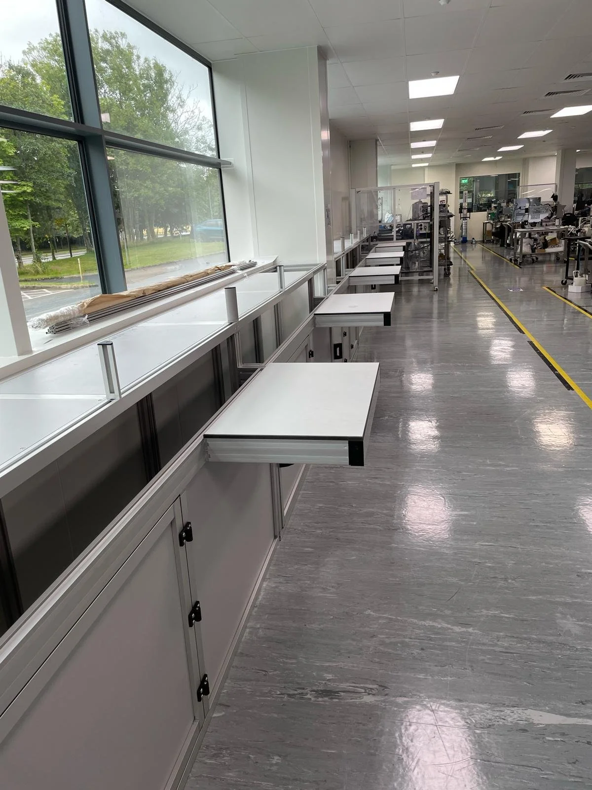 An empty manufacturing or assembly workspace with white workbenches attached to a long ledge along a large window, with a view of trees outside. The room has bright lighting, gray flooring, and appears to be under setup or renovation.