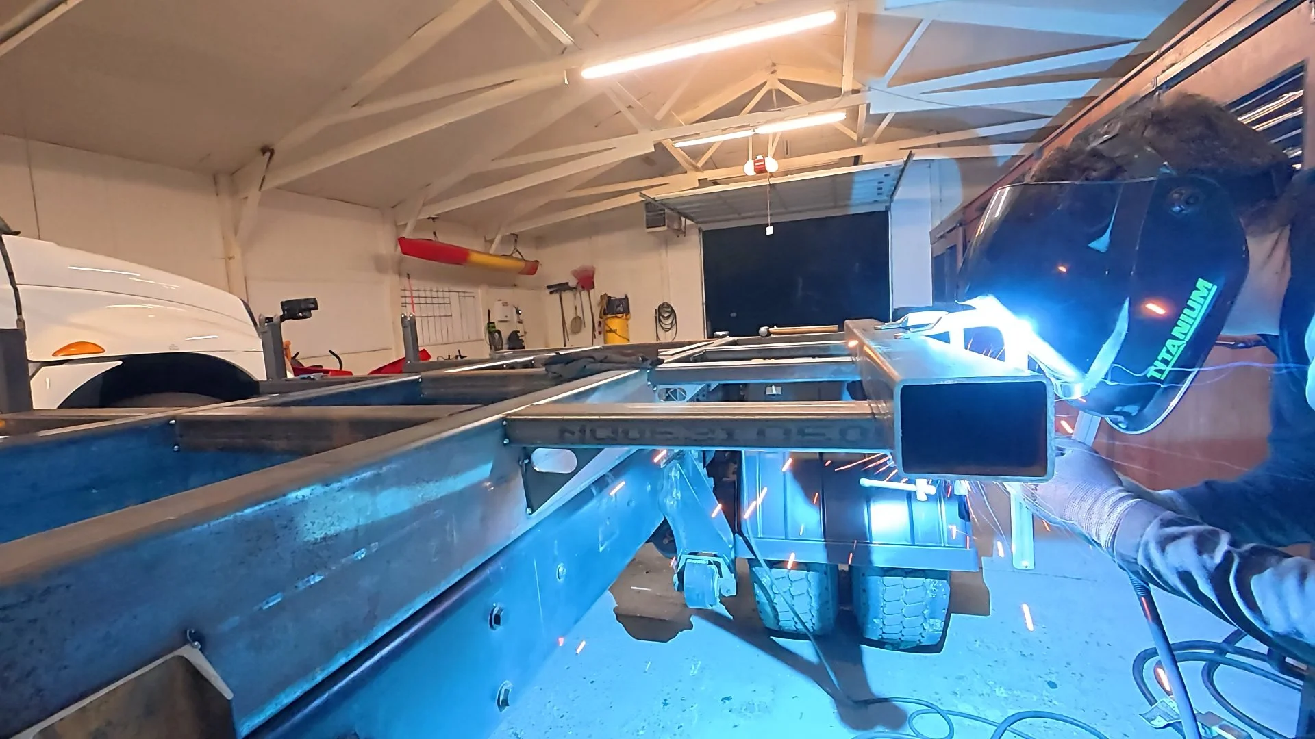 Person welding metal structure in a garage with sparks flying, wearing a dark welding helmet with a blue-green emblem, gloves, and long-sleeve shirt.