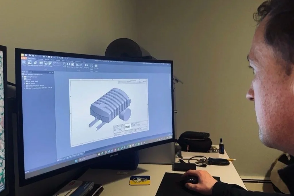 A person working on a computer displays a 3D model of an engine or mechanical part in a CAD software.