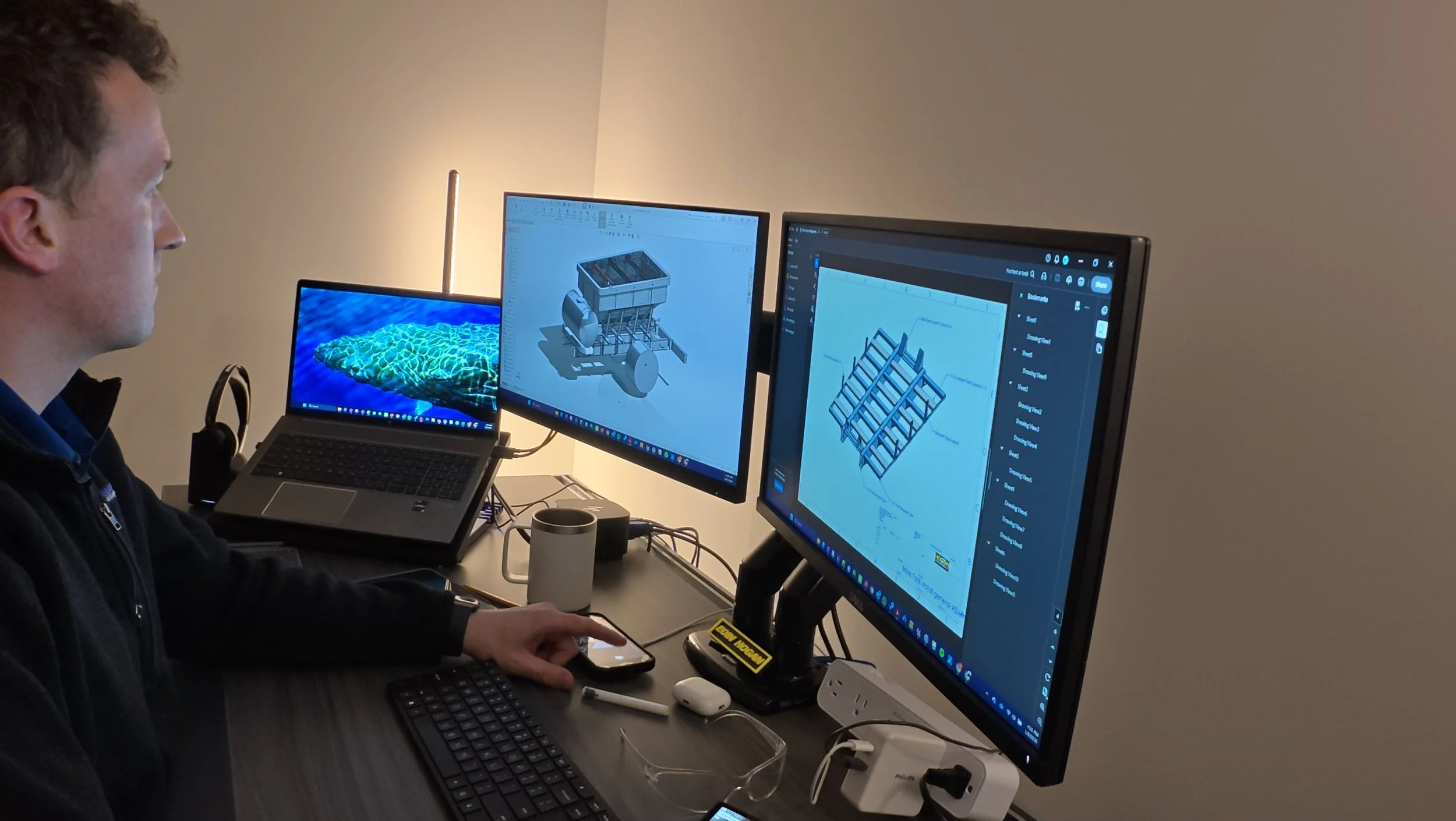 A person working at a desk with multiple monitors displaying 3D models and schematics, a laptop, a mug, safety glasses, and various electronic accessories.