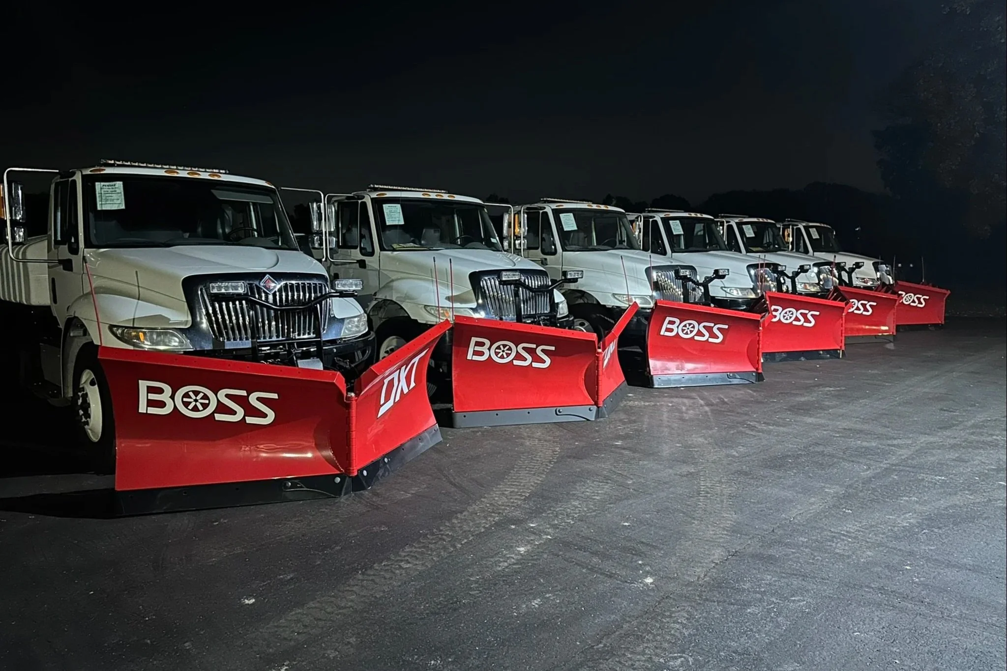 A row of large, white pickup trucks equipped with red snow plows labeled with the brand 'BOSS' parked on a dark, empty lot at night.