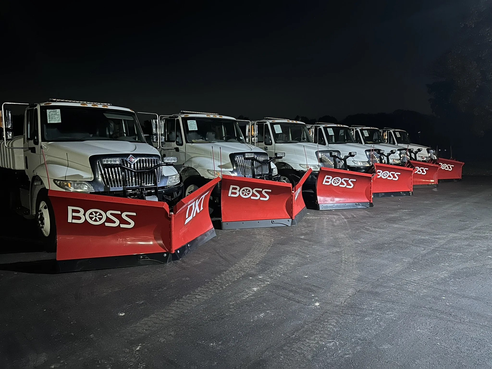 Lineup of six white utility trucks equipped with red snowplow blades marked with 'BOSS' at night on an asphalt lot.