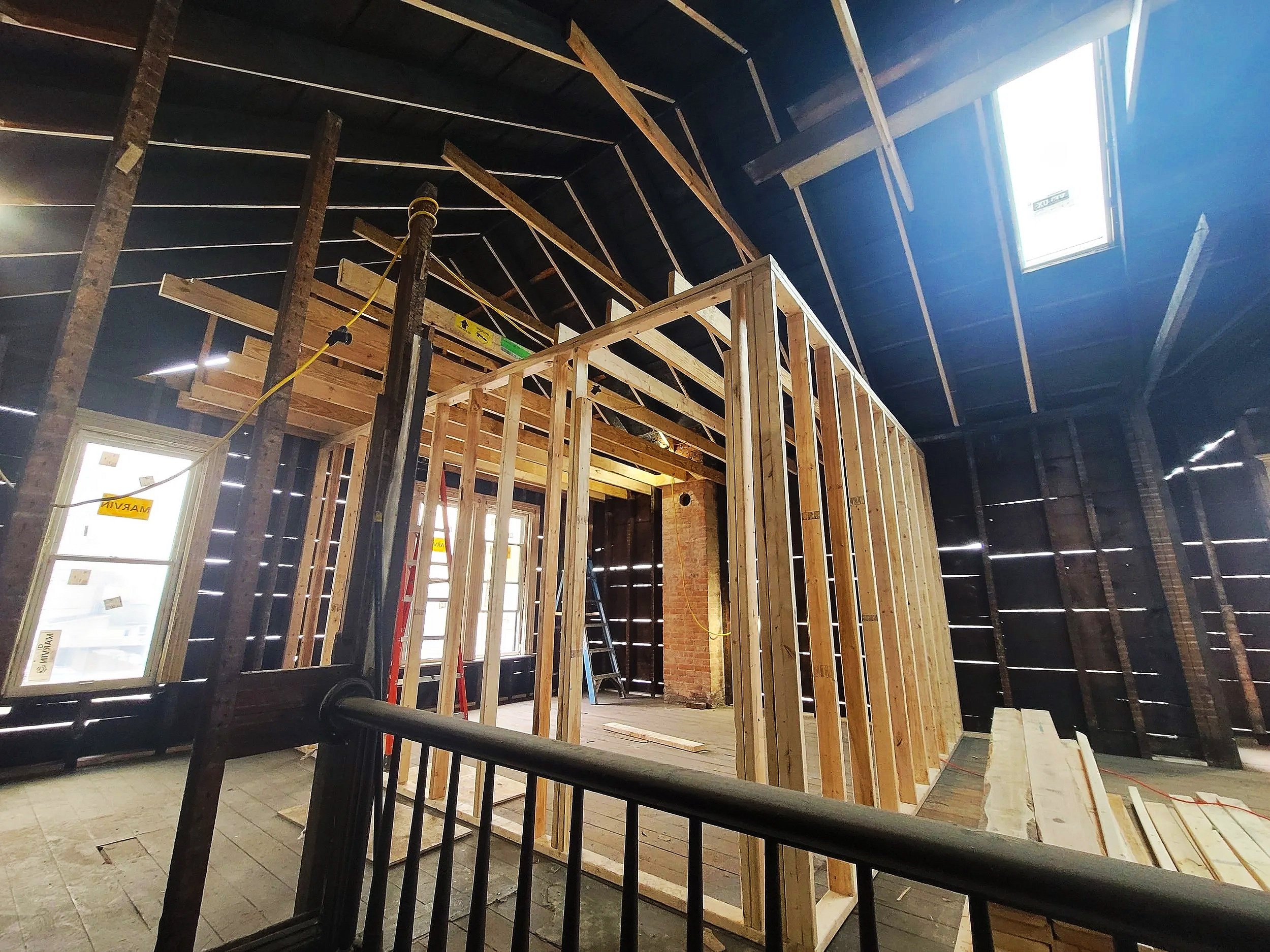 Interior of a building under construction with exposed wooden framing and support beams, windows with sunlight coming through, and construction tools visible.