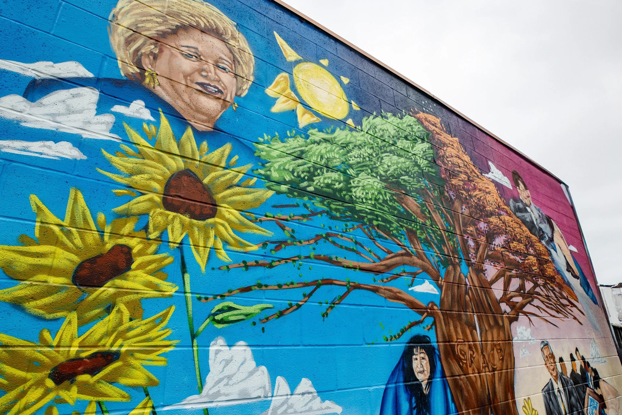 Colorful mural on a wall depicting a woman with blonde hair, sunflowers, a yellow sun, a green leafy tree, clouds, and people, set against a blue sky background.