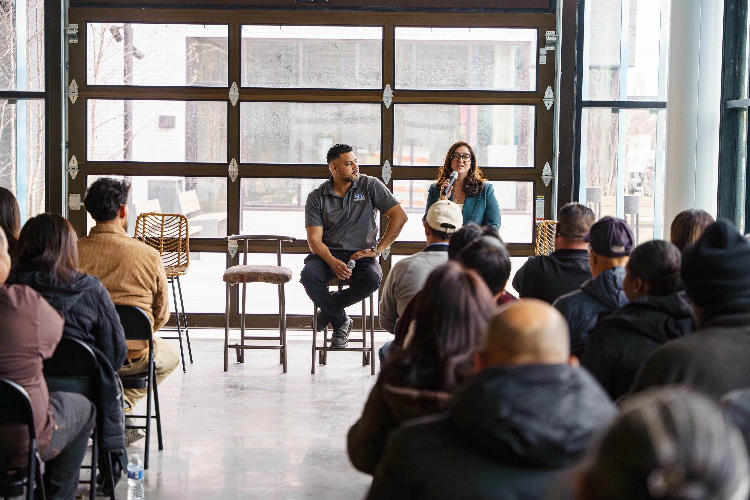 A woman speaking into a microphone while seated on a stool, with a man sitting nearby, in front of an audience in a bright room with large windows.