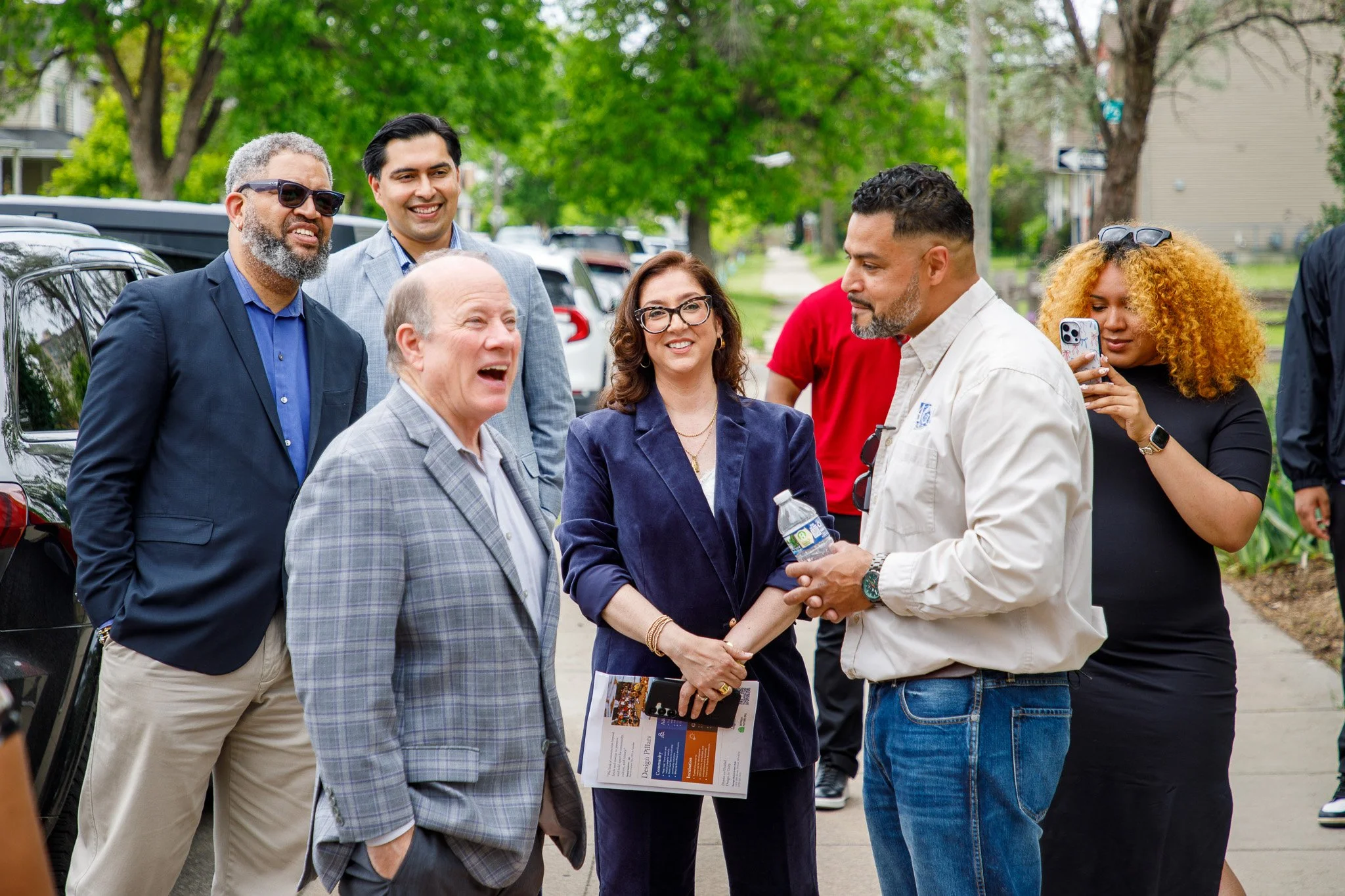 Group of diverse adults talking and smiling outdoors on a sidewalk, some taking photos, with cars parked in the background.