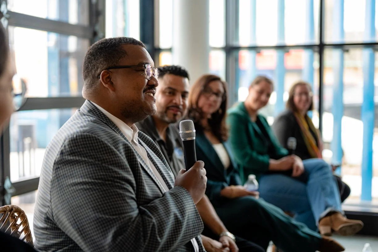 A man speaking into a microphone during a panel discussion, with several women and men sitting beside him and listening attentively in a bright, glass-walled room.