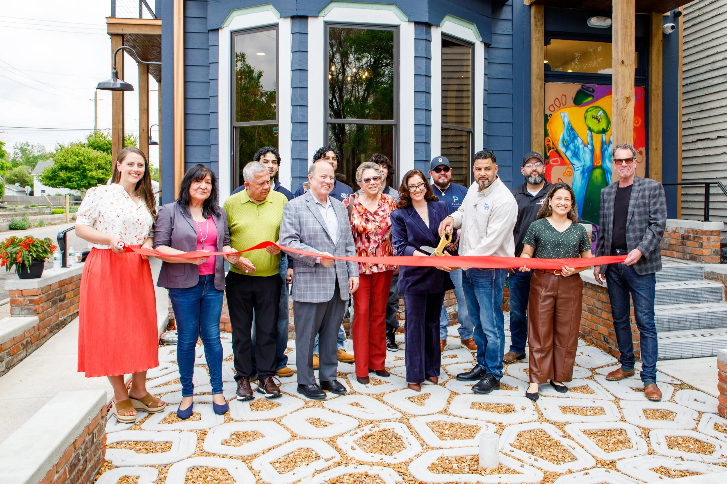 A group of people participating in a ribbon-cutting ceremony outside a building with a blue exterior and large windows. The group includes men and women of various ages, some holding a large pair of scissors. The setting is decorated with potted plants and artwork on the wall.