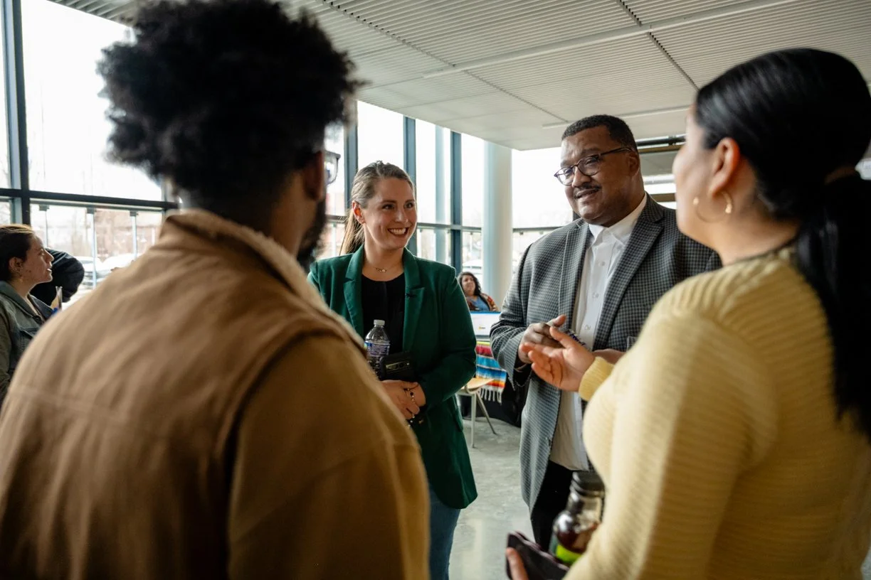 Four diverse people are engaged in a conversation inside a modern building with large windows, with a woman in a green blazer smiling at a man in glasses and a gray checkered blazer, while two other women listen.