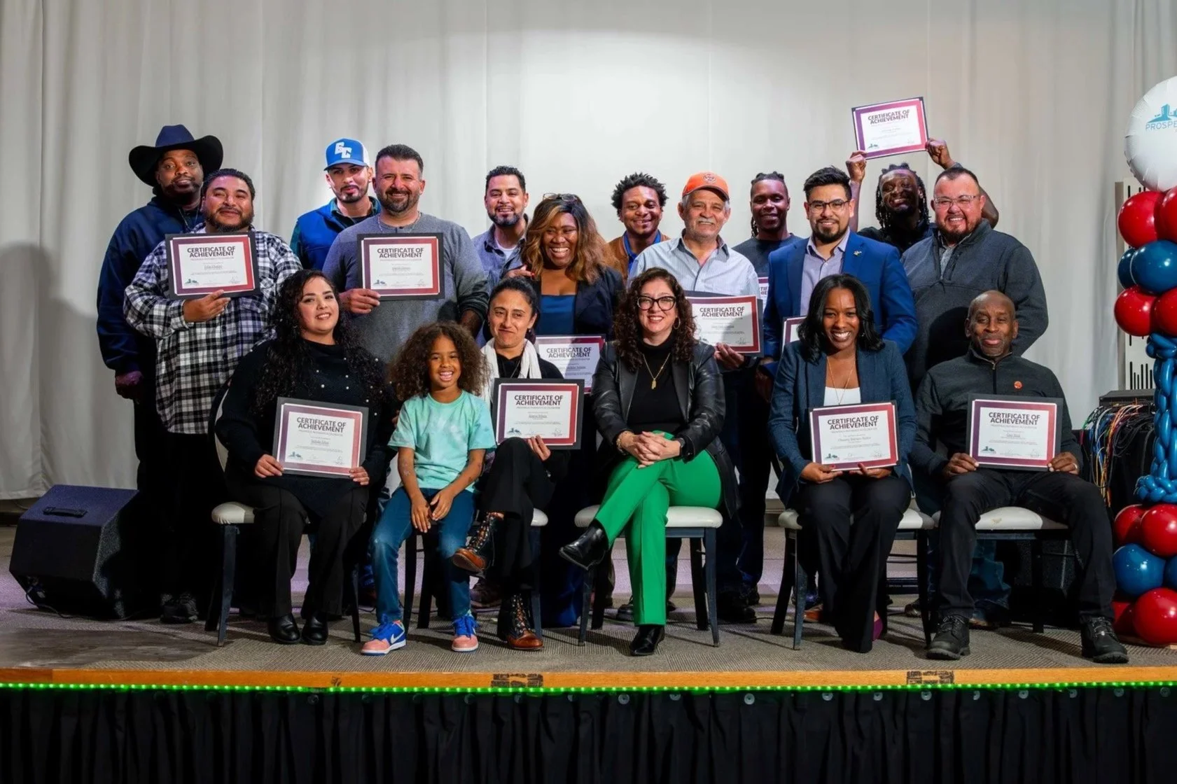 Group of diverse people on stage holding certificates, some sitting, some standing, celebrating achievement, with celebratory balloons on the right.