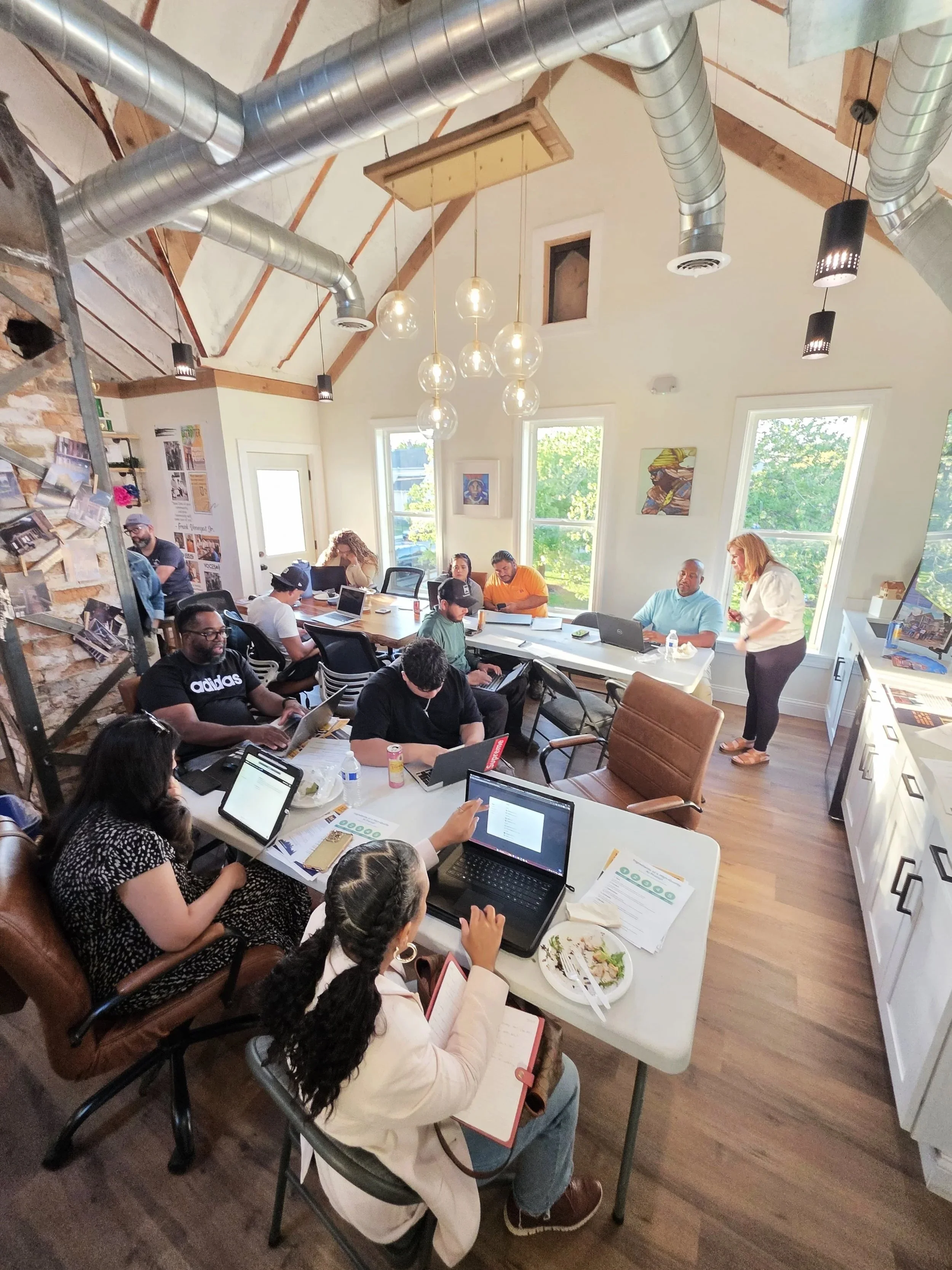 A group of people in a bright, modern meeting room with large windows, working on laptops and tablets, some sitting at a split white and wood table, with a woman standing near the white cabinetry at the back.