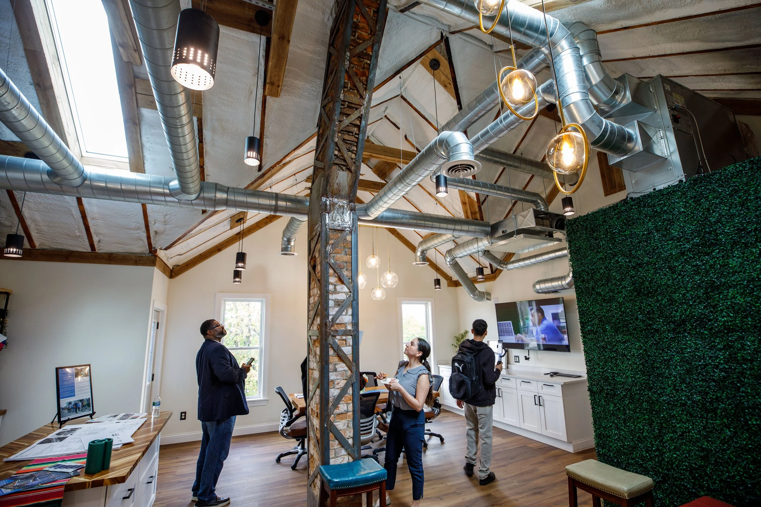 People in a modern office space with a high, angled ceiling, exposed ductwork, large windows, and a green wall. The office has wooden accents, a conference table, chairs, and a wall-mounted television.
