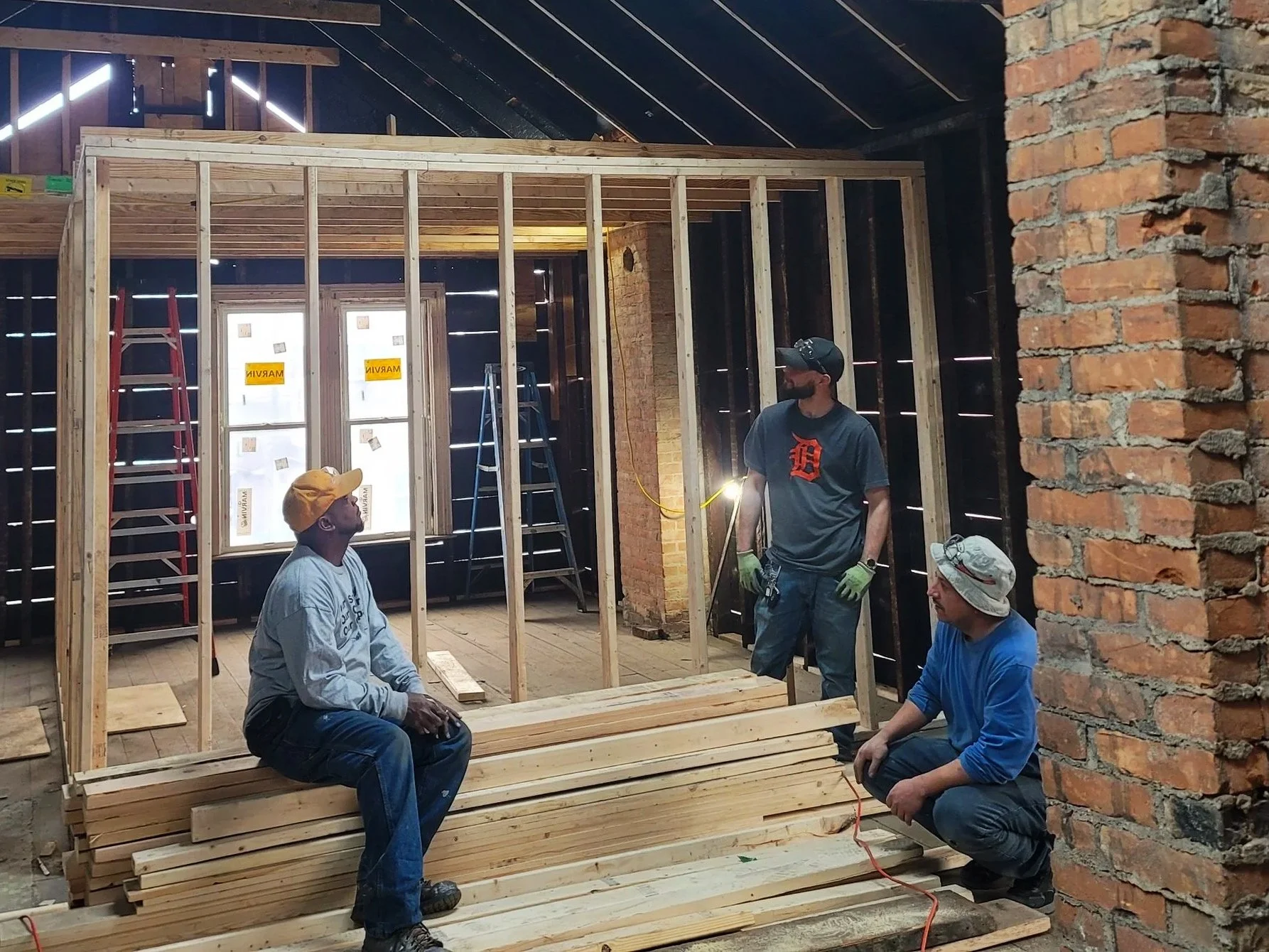 Three men working on framing a wall in a construction site, with wooden studs, ladders, and a brick wall in the background.