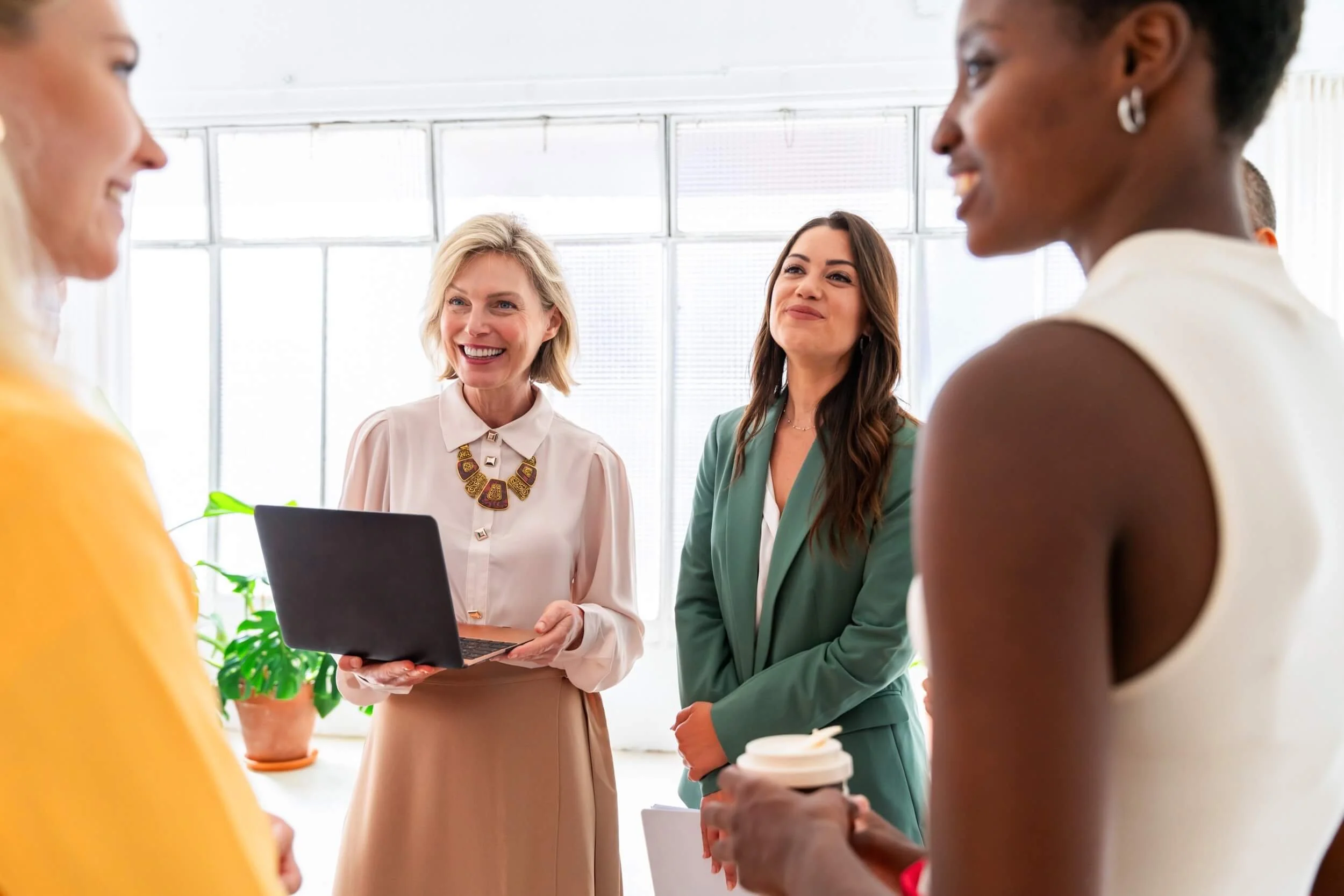 A group of women having a professional meeting or discussion in a bright office space. The woman in the center is holding a laptop and is smiling, while others are listening and engaging.