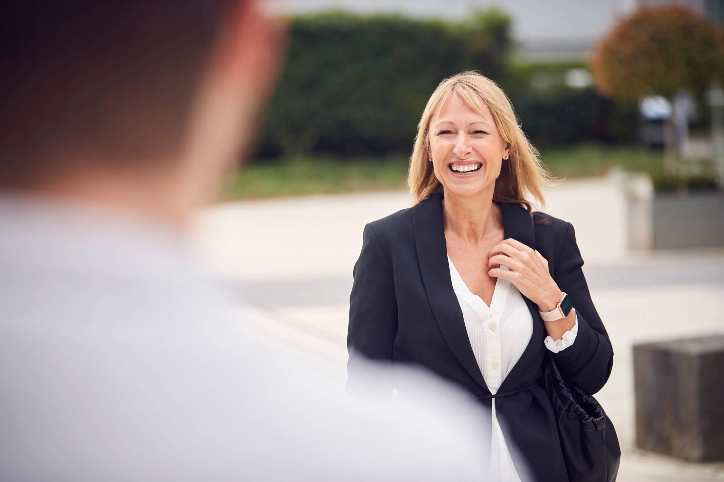 A woman with blonde hair smiling and talking outdoors, wearing a black blazer and white shirt.