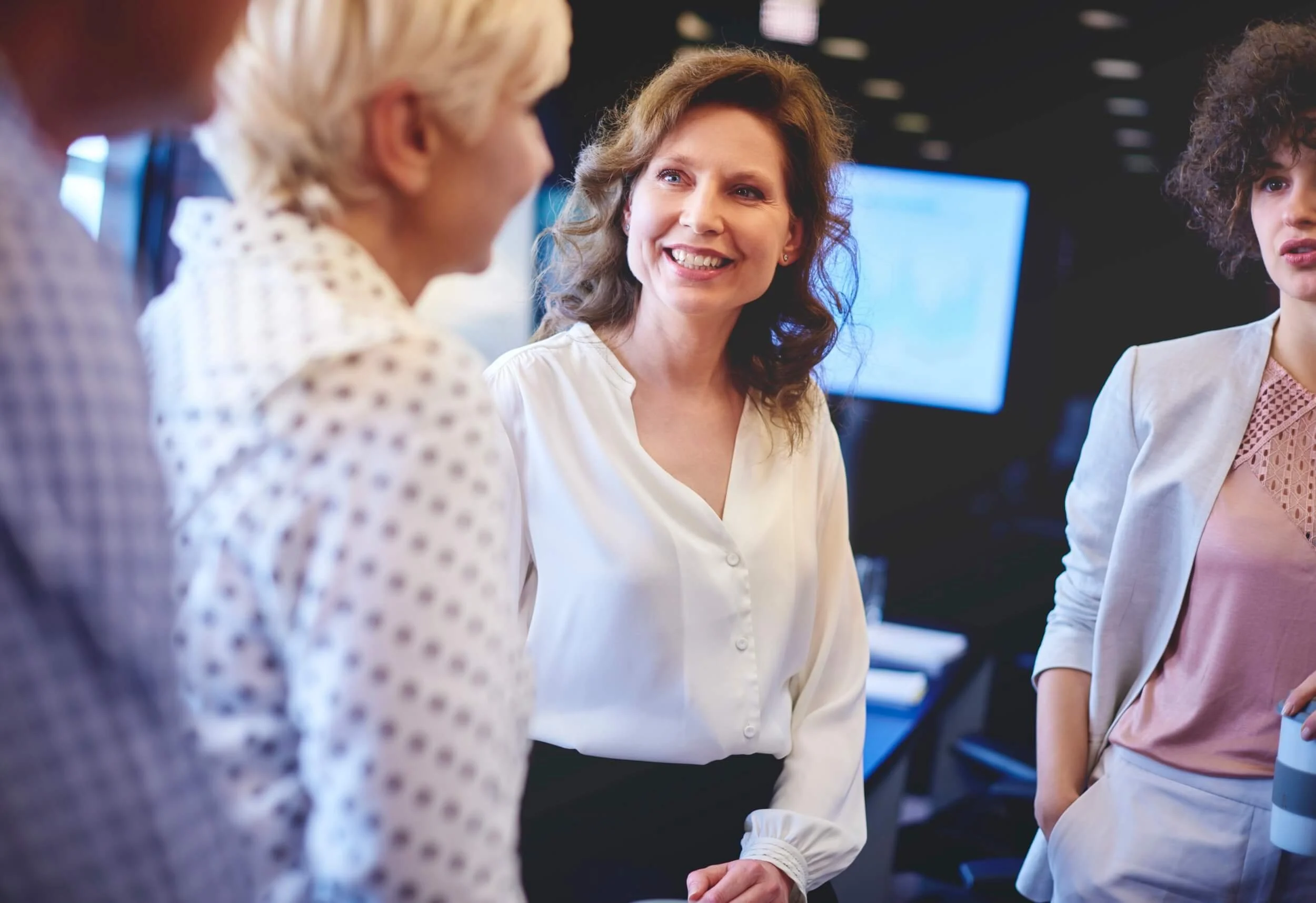 Four women engaged in conversation in a professional setting, with a large monitor in the background.