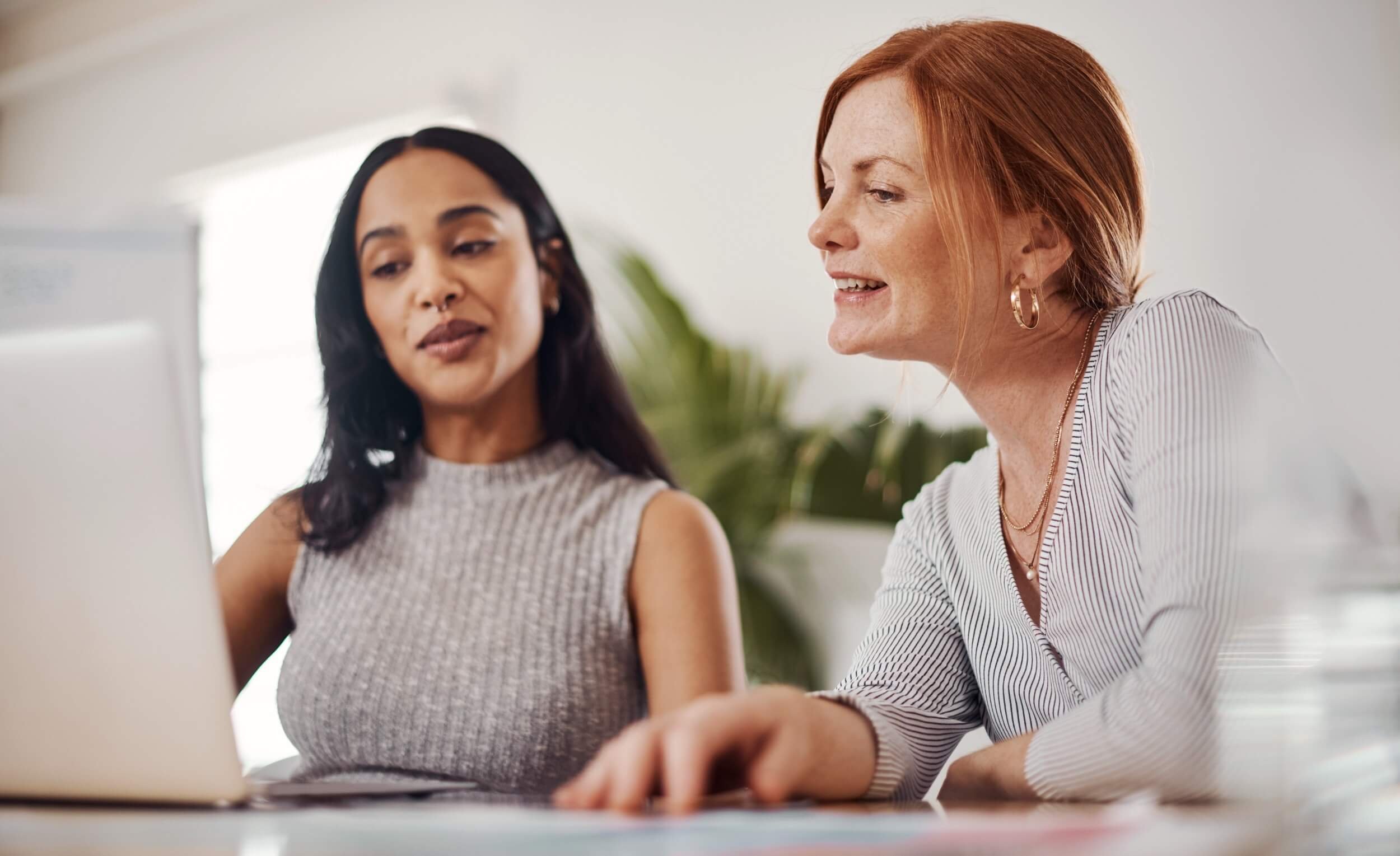 Two women working together at a desk, looking at a laptop screen, in an office setting.