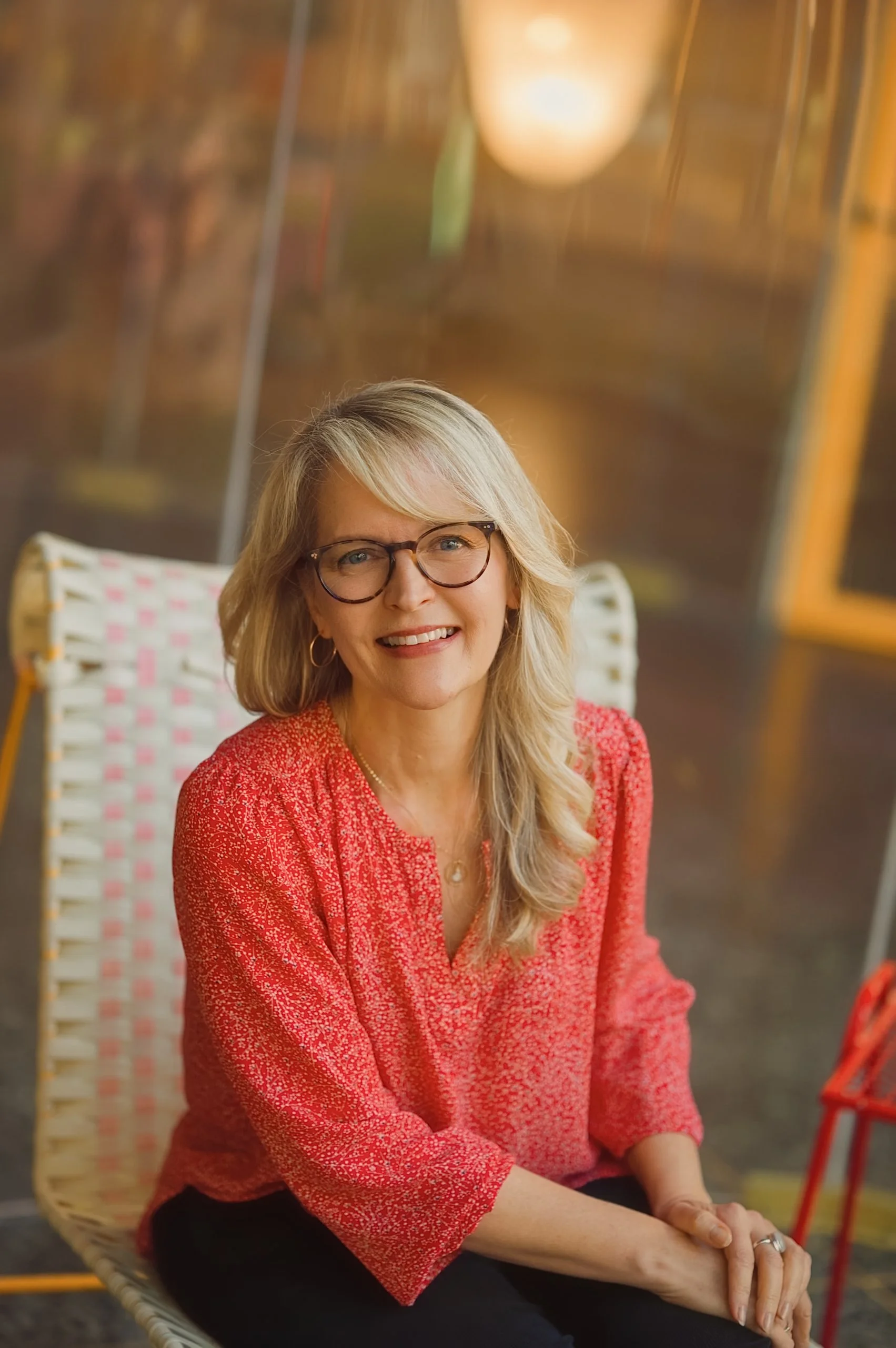 A smiling middle-aged woman with blonde hair, wearing glasses and a red patterned blouse, is sitting on a woven chair in a warmly lit room.