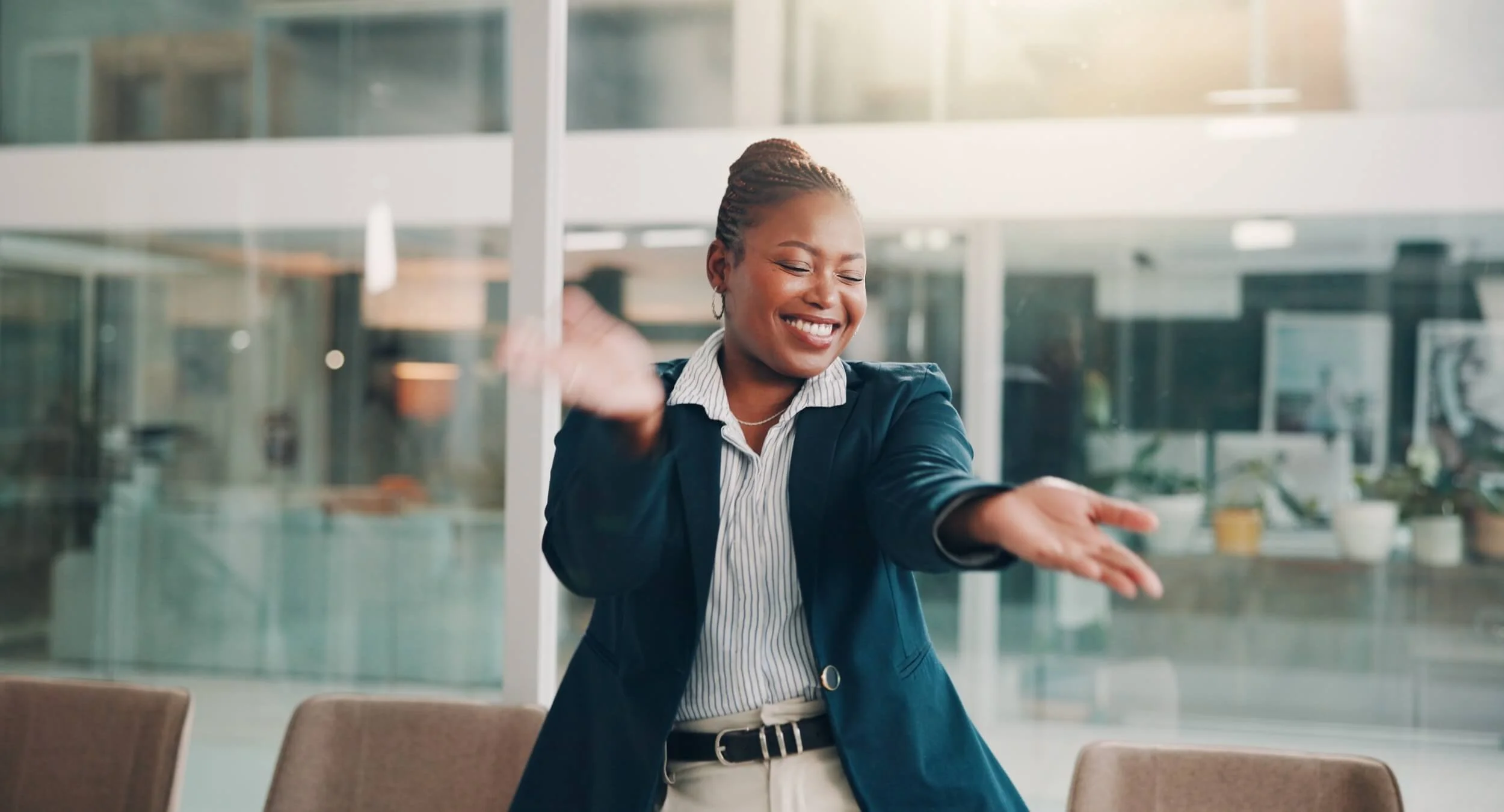 A smiling woman in business casual attire dancing or stretching in a modern office with glass walls.