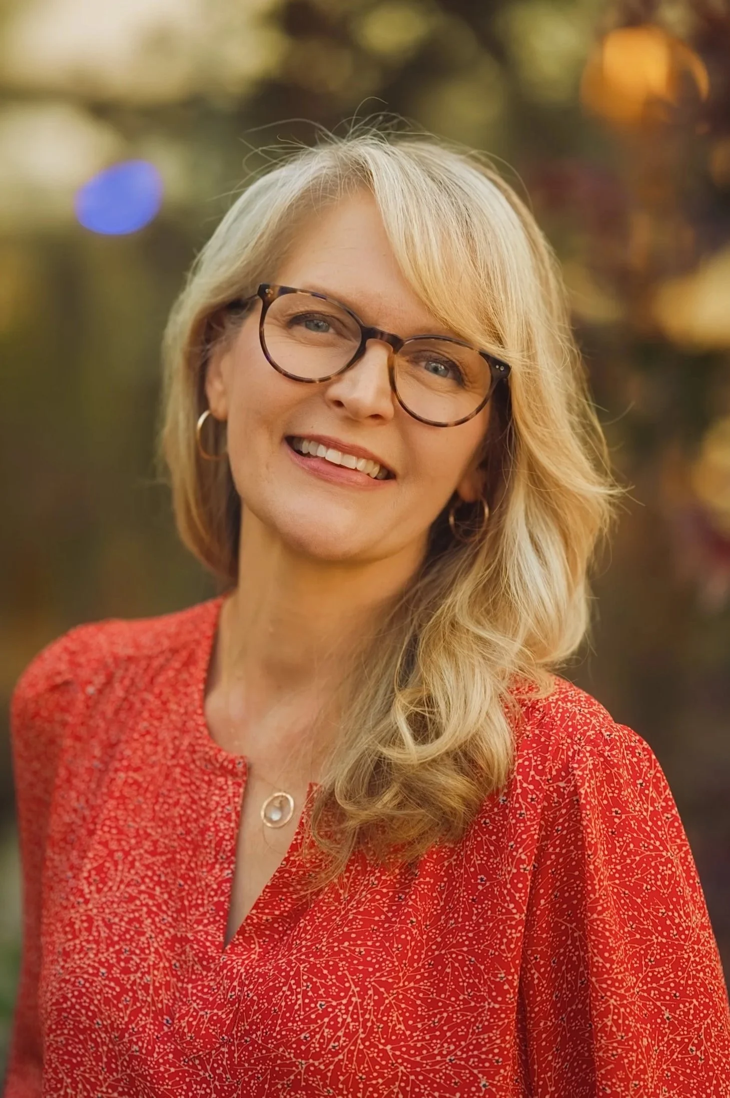 A smiling middle-aged woman with blonde hair, glasses, and hoop earrings, wearing a red patterned blouse, outdoors with a blurred background of trees and lights.