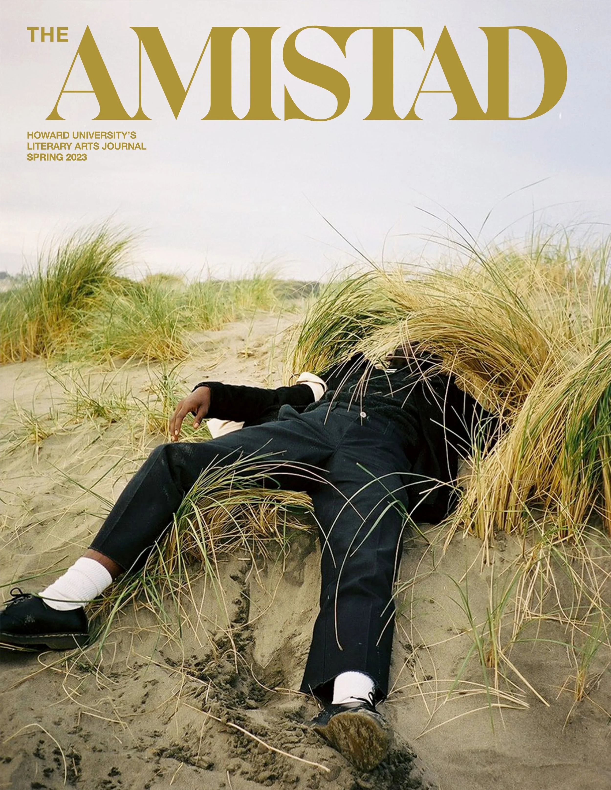Cover of The Amistad, Howard University’s literary arts journal spring 2023, featuring a person lying on sand dunes with tall grass, dressed in dark clothing and white socks, with their face obscured by tall grass.