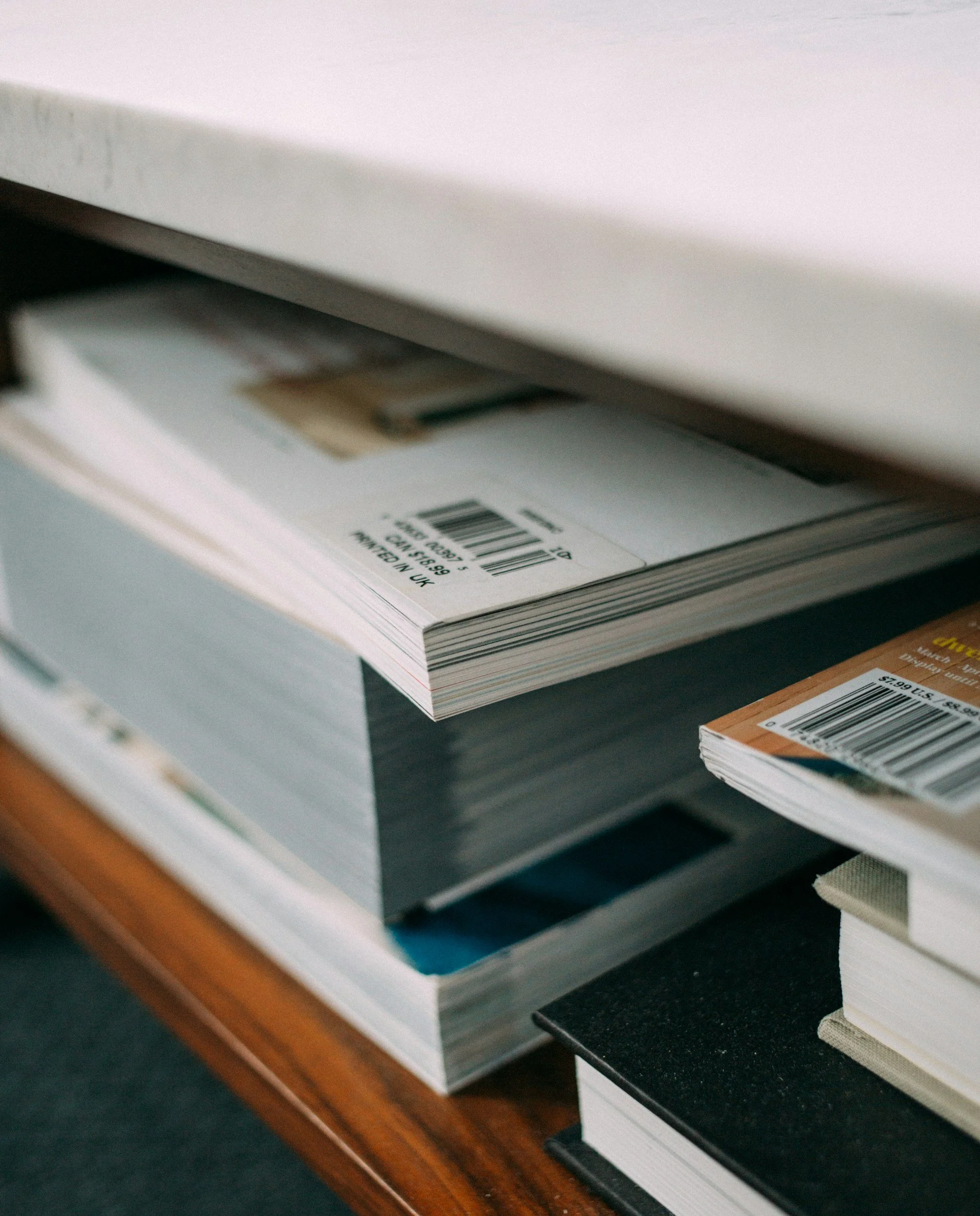 Stack of magazines and books on a wooden desk, with barcode labels visible on some.