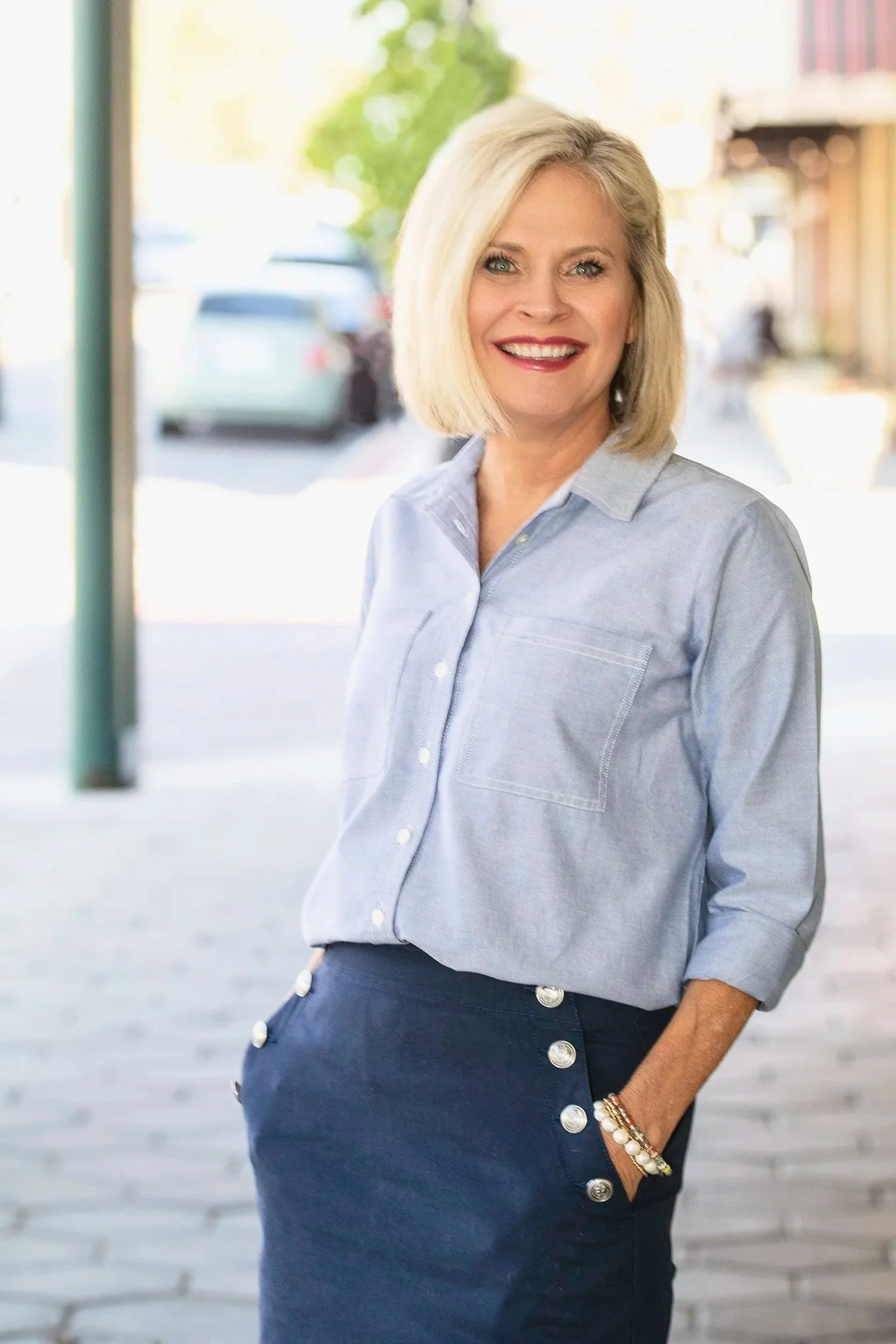 A woman with shoulder-length blonde hair smiling outdoors, wearing a light blue button-up shirt and dark blue skirt with large buttons, standing on a paved sidewalk.