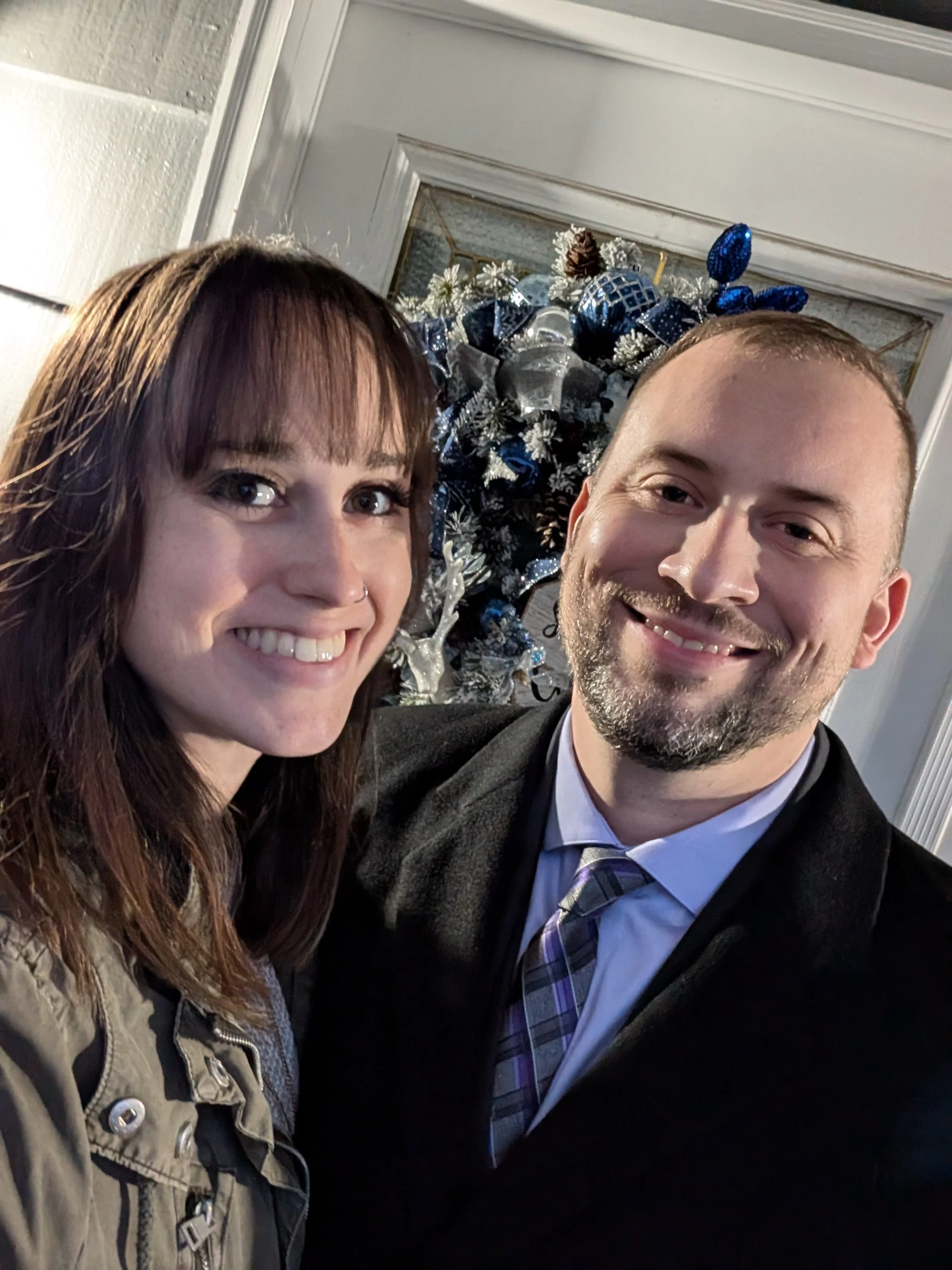 A smiling woman with brown hair and a man with a beard and short hair taking a selfie in front of a Christmas wreath decorated with blue, silver, and white ornaments.