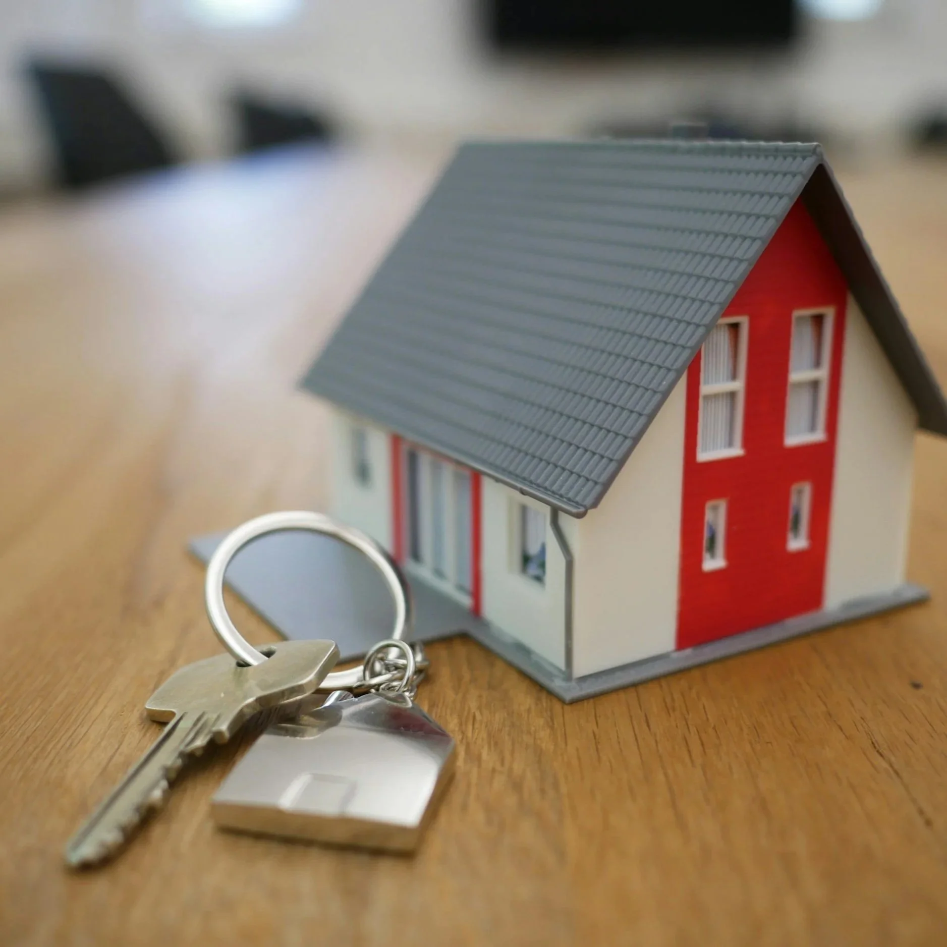 Model house with grey roof and red and cream walls, placed on a wooden table, with a key and keychain in the foreground.