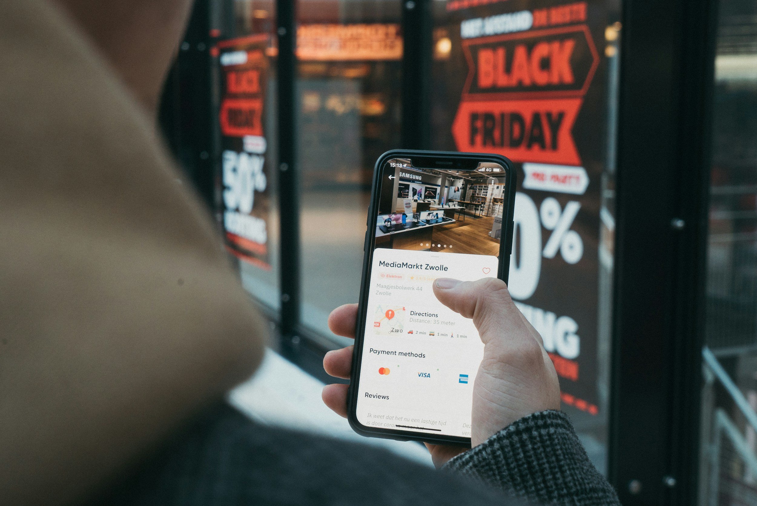 A person holds a smartphone displaying details of MediaMarkt Zwolle, an electronics store, in front of a large black and red Black Friday sale sign outside the store.